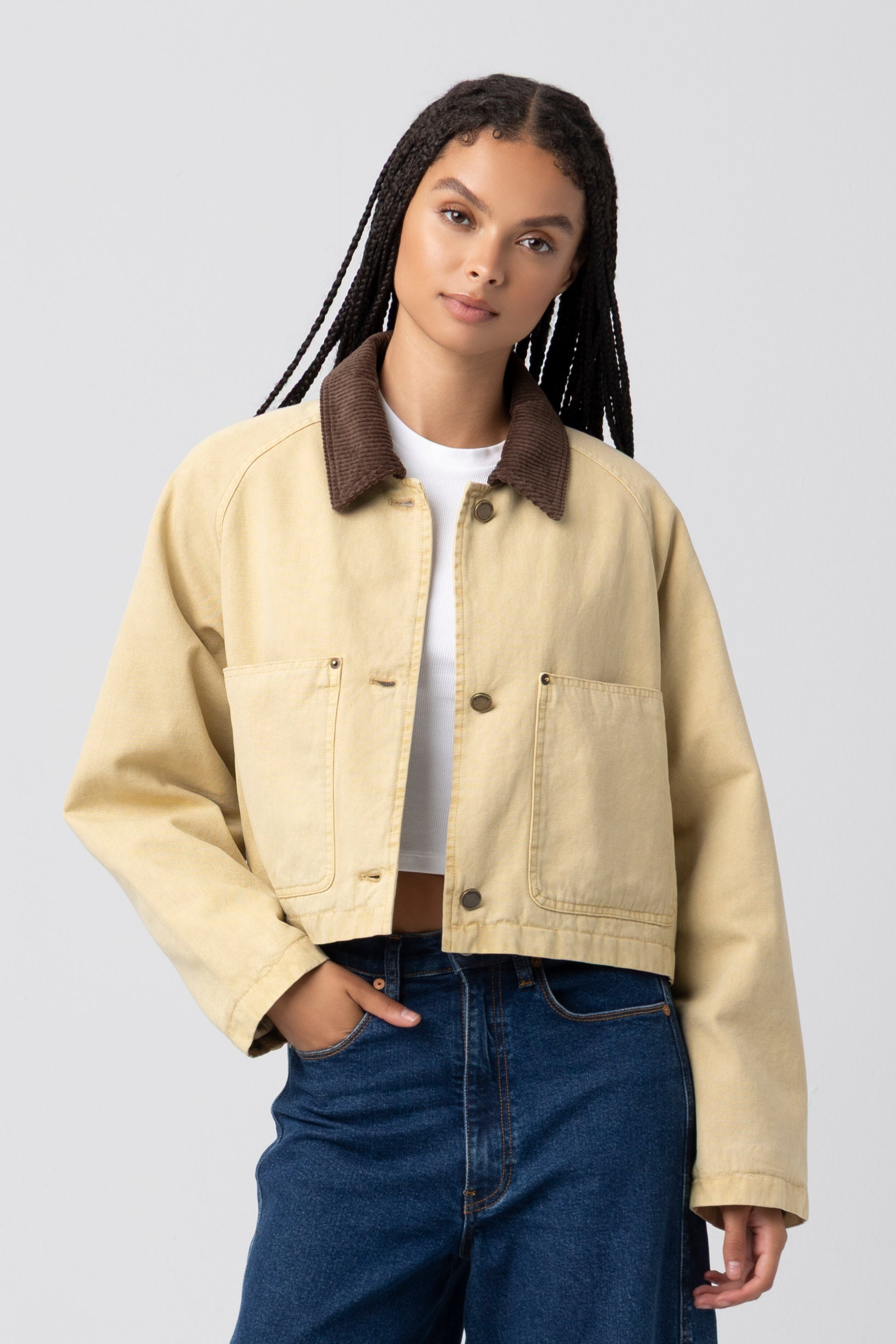 A woman with long braids wears the Breaking Camp Barn Jacket by Blank, a cropped beige jacket with a dark brown collar, relaxed fit, and durable cotton shell, layered over a white shirt and blue jeans as she stands against a plain light gray background.
