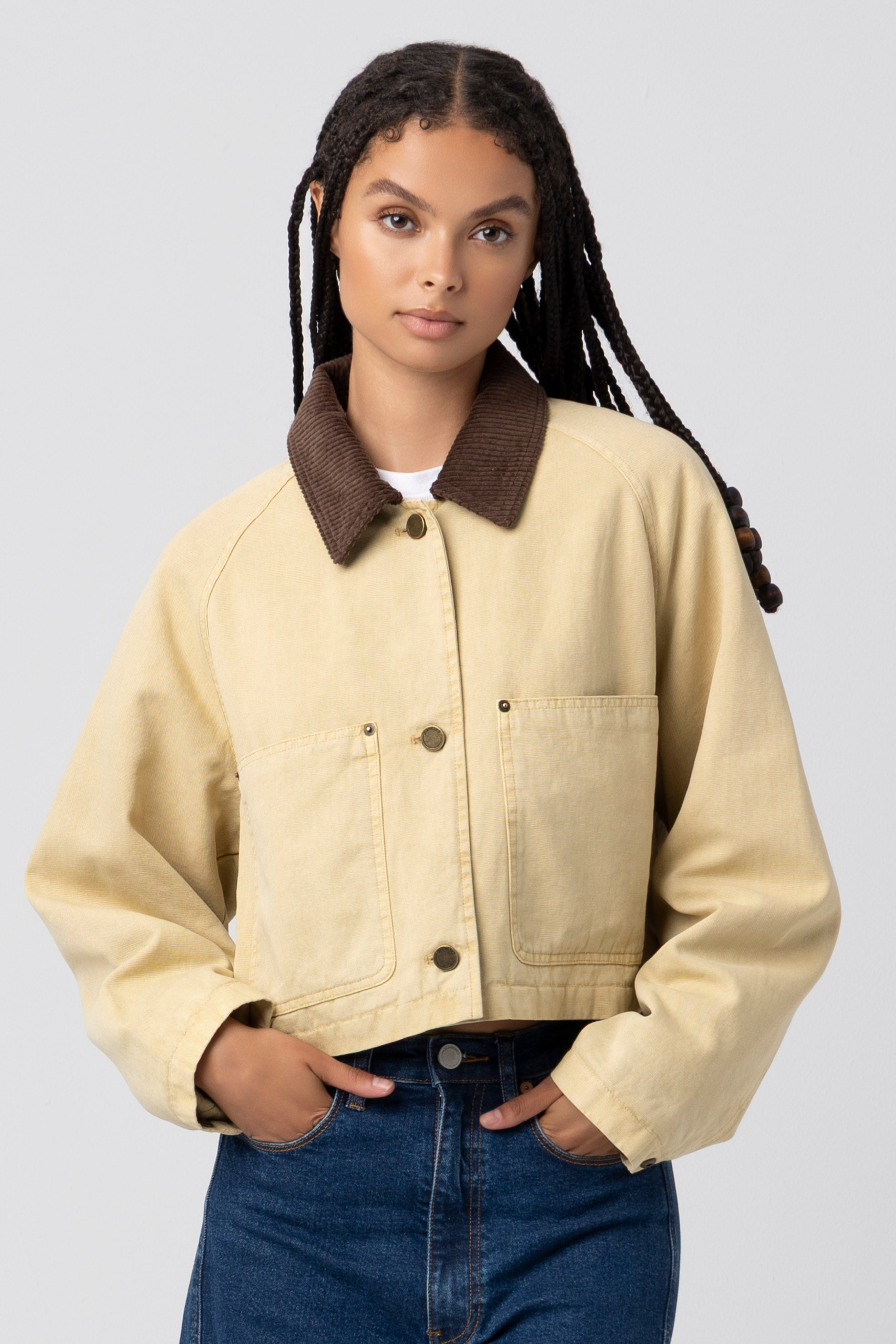 A woman with long, braided hair stands against a plain light gray background wearing Blank’s Breaking Camp Barn Jacket in GOLD KHAKI—a light beige cropped style featuring a dark brown collar and a durable cotton shell with a relaxed fit for comfort and style—paired with blue jeans.