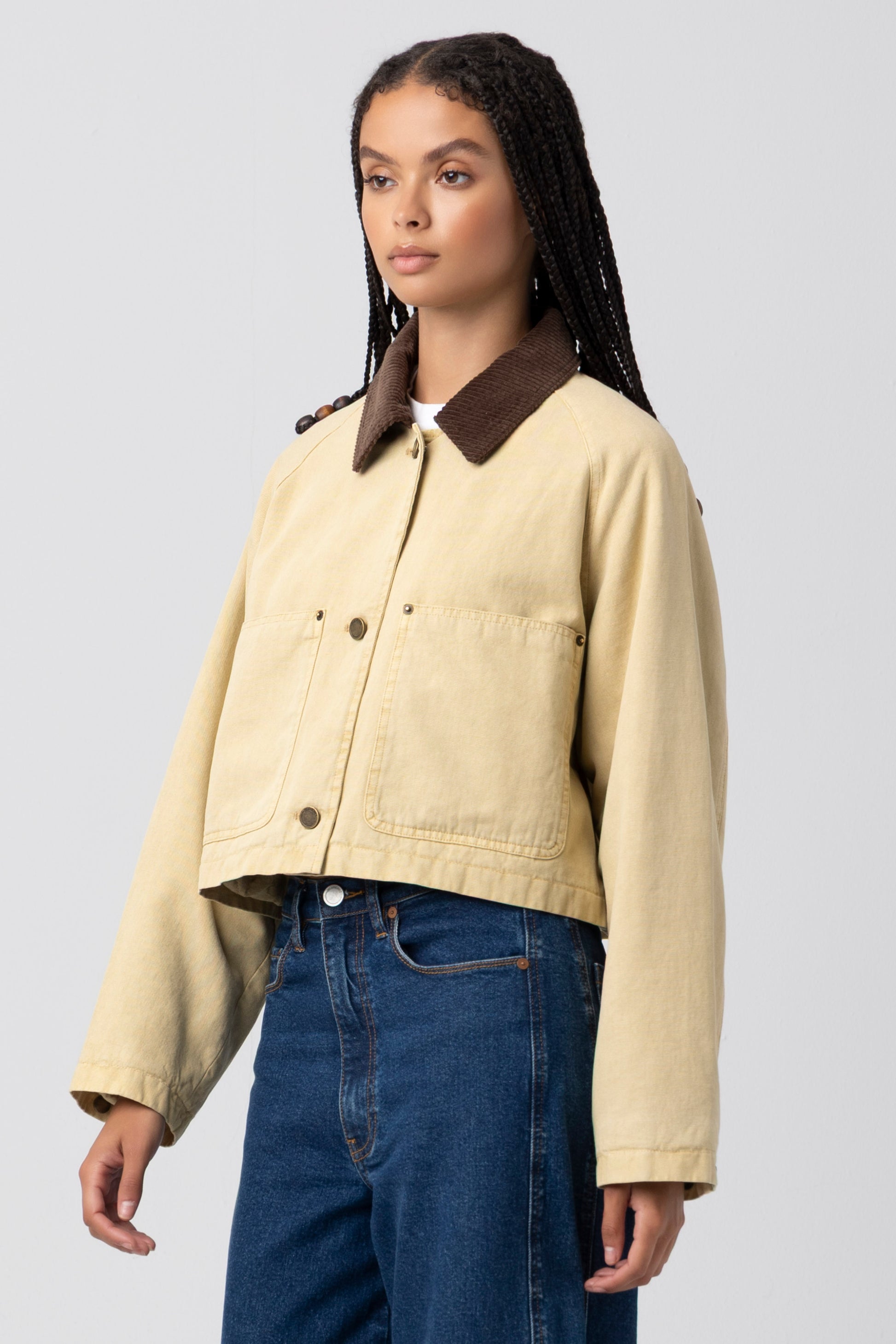 A woman with long braided hair wears the Breaking Camp Barn Jacket by Blank—a beige jacket with a durable cotton shell, large front pockets, and a brown collar—paired with blue jeans. She stands facing slightly to the side against a plain light background.