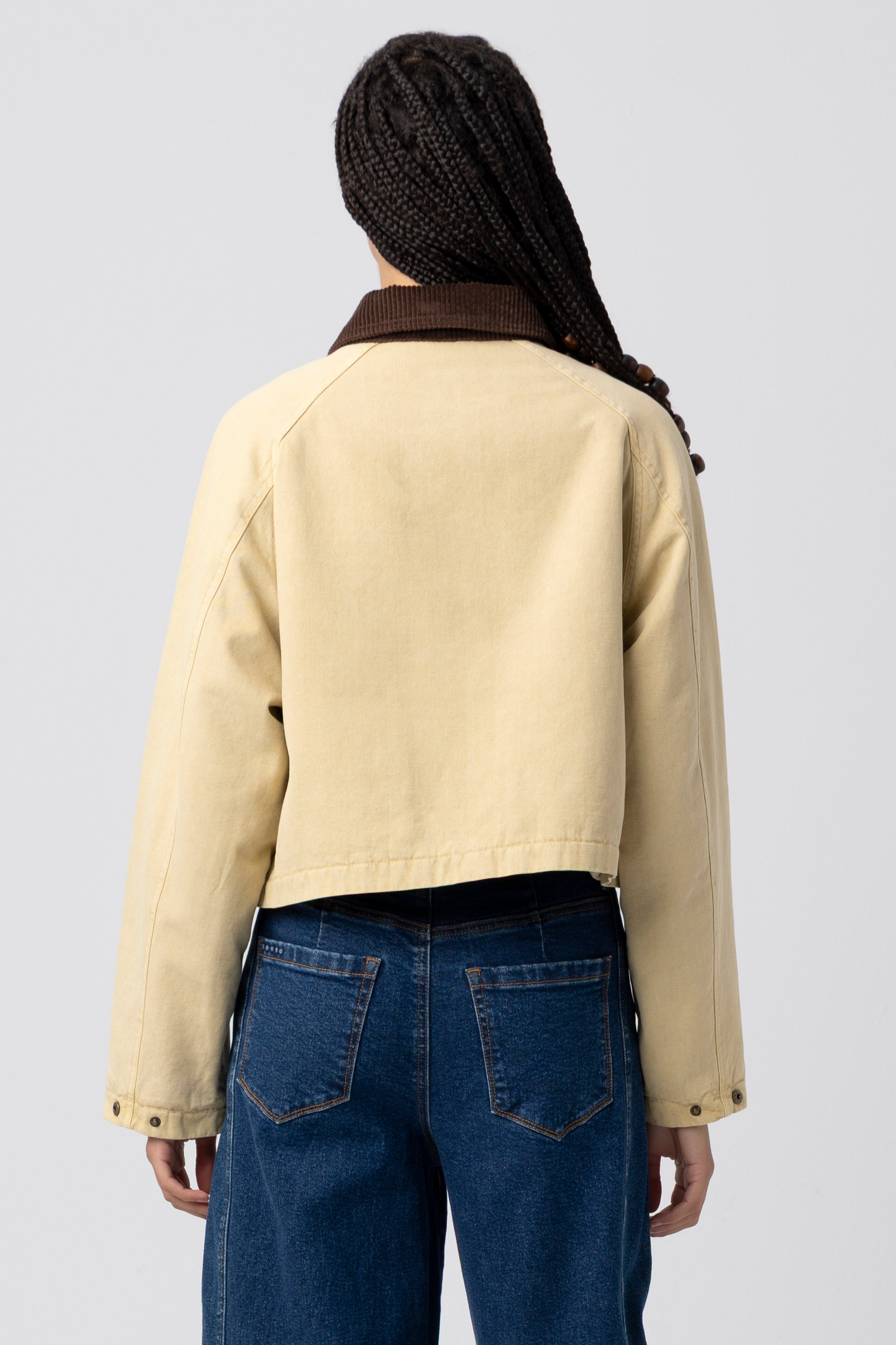 A person with braided hair is shown from the back, wearing the Blank Breaking Camp Barn Jacket—a light yellow jacket with a dark brown collar and a relaxed fit, crafted from a durable cotton shell for comfort and style—paired with blue jeans.