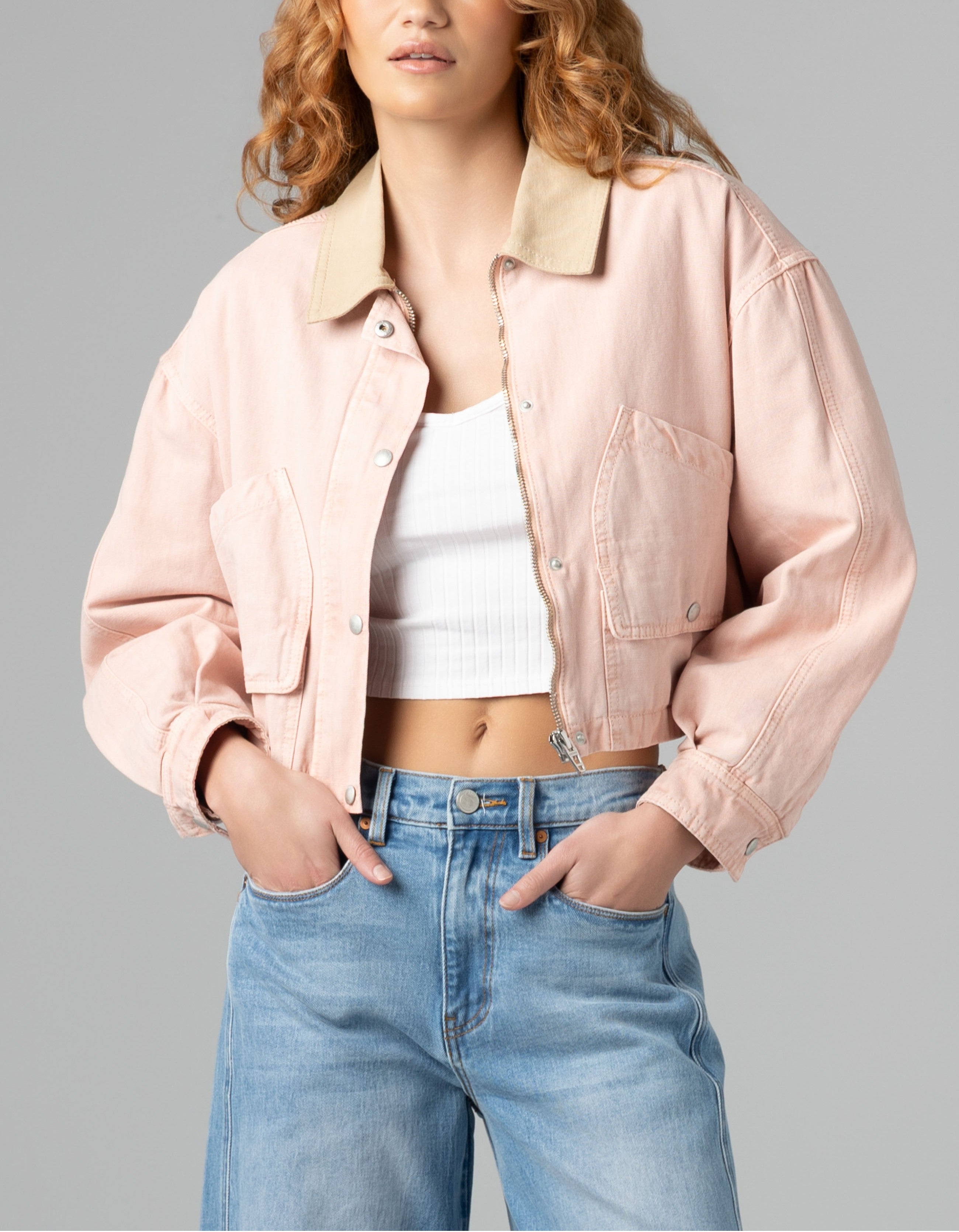 A person wearing the Blank Lawn Party Barn Jacket in light pink over a white ribbed crop top and blue high-waisted jeans stands with hands in the jacket pockets against a plain gray background.