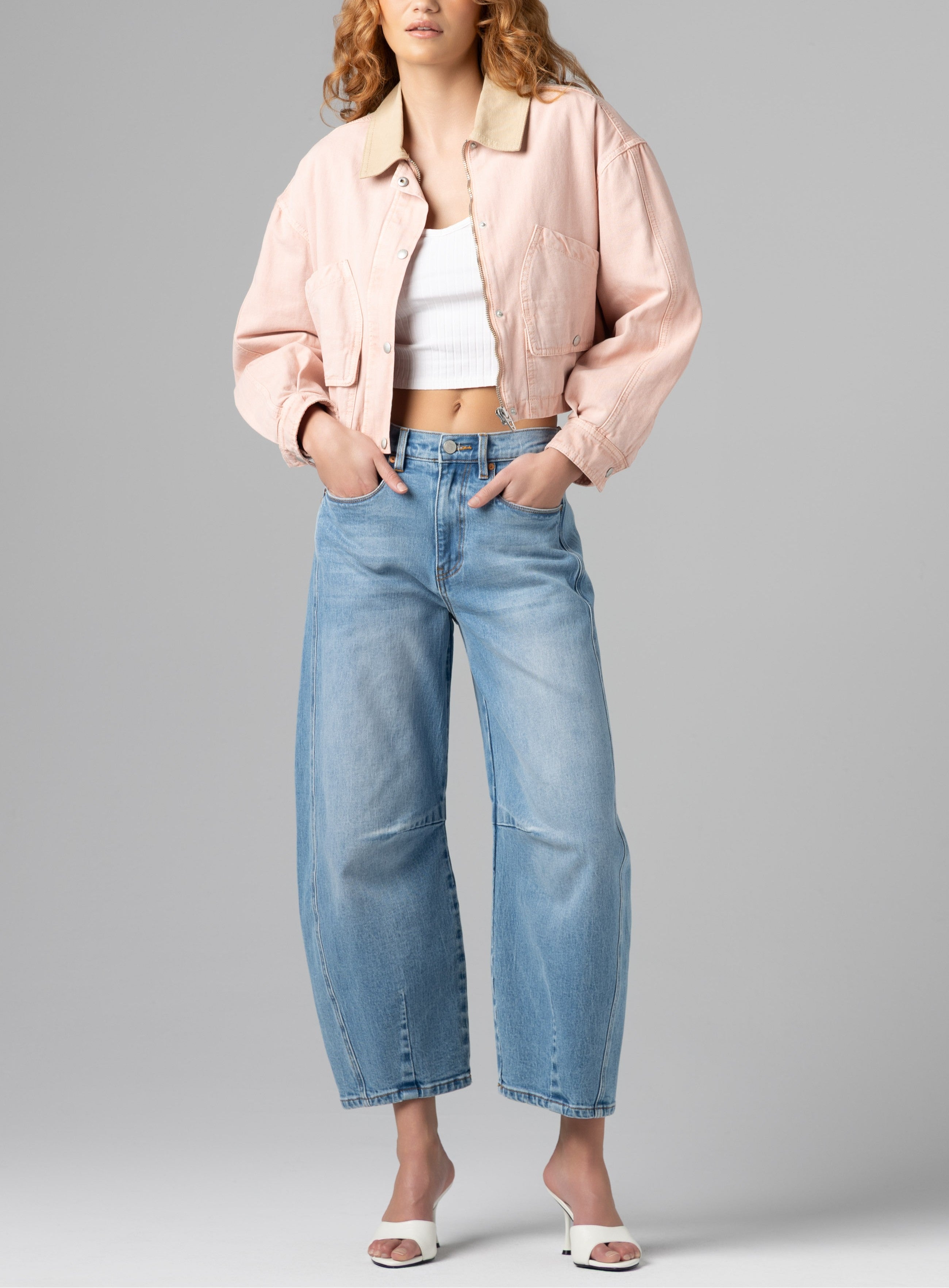 Wearing the Lawn Party Barn Jacket by Blank, a person pairs it with a white crop top, light blue wide-leg jeans, and white open-toe heels, standing hands-in-pockets against a plain gray background.