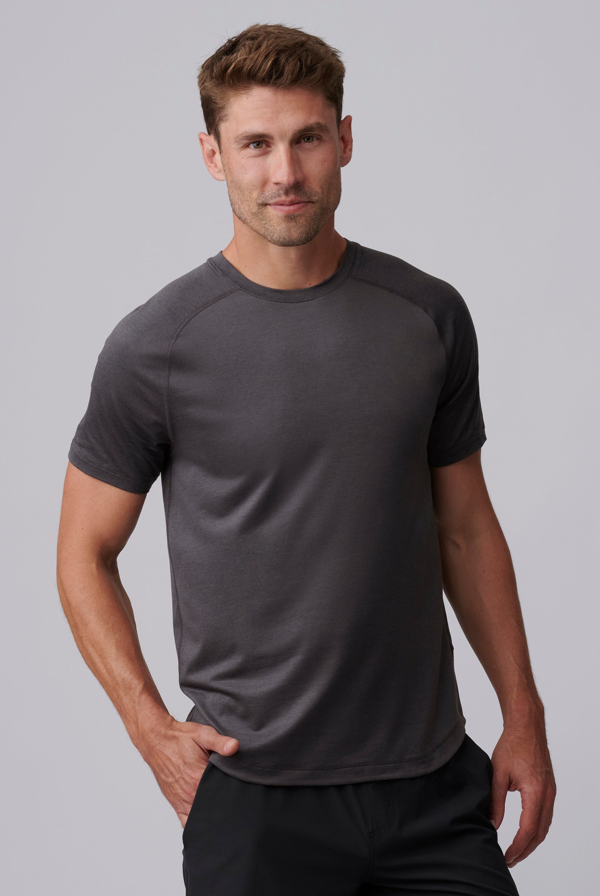 A man with short brown hair wears the South Moon Under Atmosphere Tee in Dusty Sage Heather and black pants, standing against a light gray background. He looks at the camera with a slight smile, one hand in his pocket.