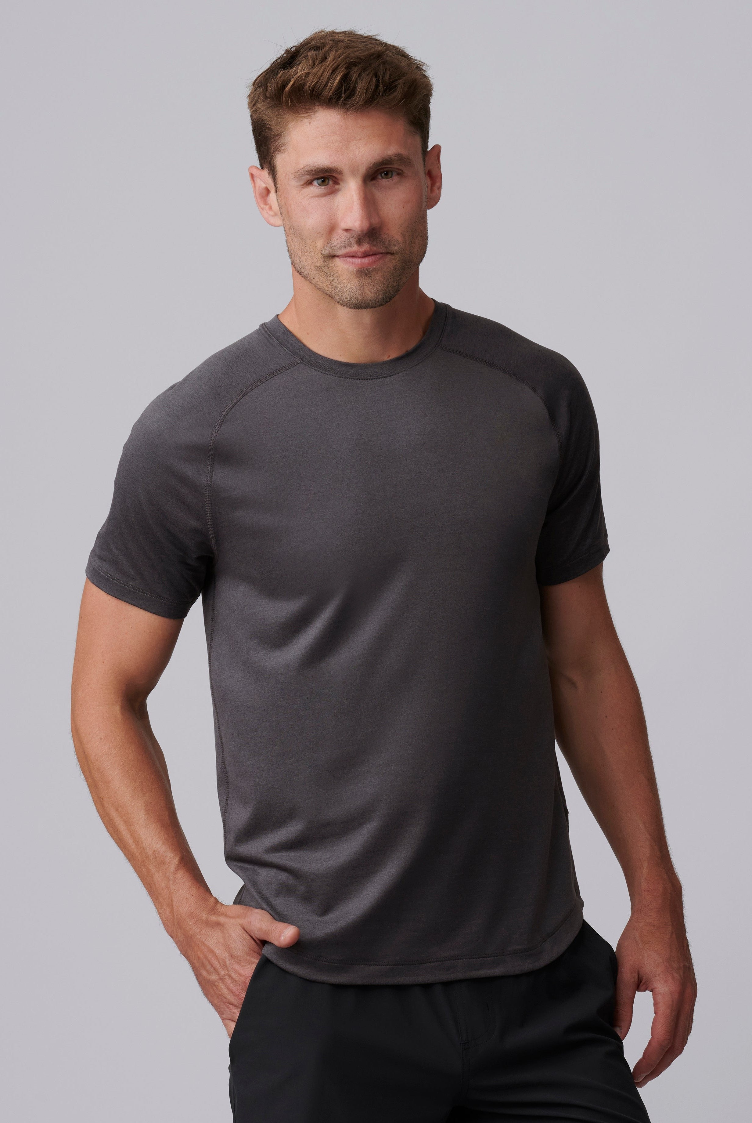 A man with short brown hair wears the South Moon Under Atmosphere Tee in Dusty Sage Heather and black pants, standing against a light gray background. He looks at the camera with a slight smile, one hand in his pocket.