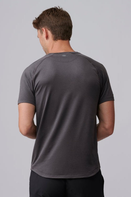 A man faces away from the camera wearing the South Moon Under Atmosphere Tee, a short-sleeved, dark gray lightweight performance T-shirt, along with black pants against a plain light gray background.