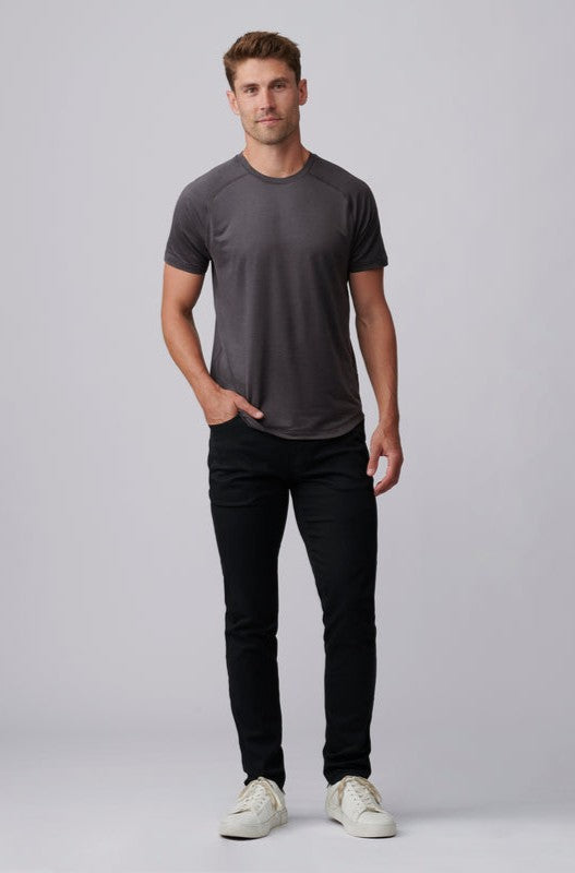 A man stands against a plain light gray background, wearing the South Moon Under Atmosphere Tee in dark gray, paired with black jeans and white sneakers. He has one hand in his pocket and maintains a neutral expression.