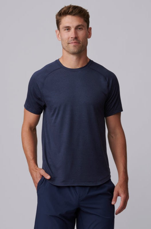A man with short brown hair stands against a light gray background, wearing the South Moon Under Atmosphere Tee in NAVY HEATHER with matching shorts, one hand in his pocket and a slight smile.