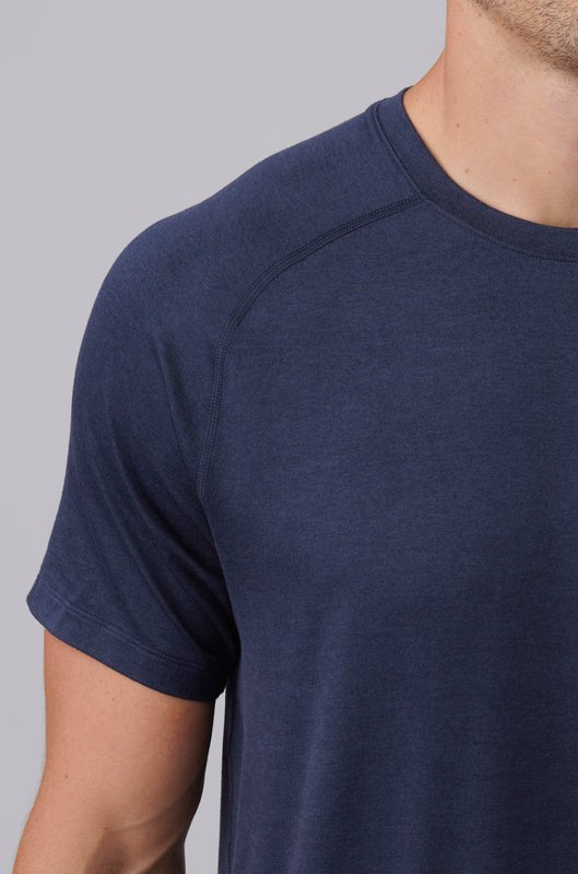 A close-up shows a person wearing the Atmosphere Tee by South Moon Under—a short-sleeved, dark blue crew neck with a classic fit—against a plain light gray background; only their upper torso and part of their face are visible.