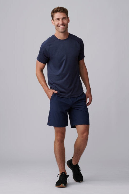 A man smiles with one hand in his pocket, wearing the Atmosphere Tee by South Moon Under in navy, paired with matching navy shorts and black sneakers, standing against a plain gray background.