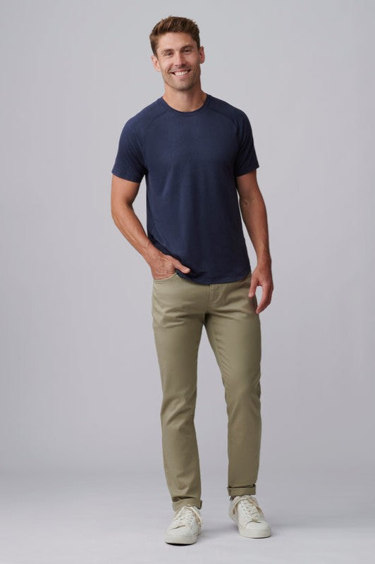 A man stands smiling against a plain gray background, wearing the navy blue Atmosphere Tee by South Moon Under, paired with khaki pants and white sneakers. One hand is in his pocket while the other rests at his side.