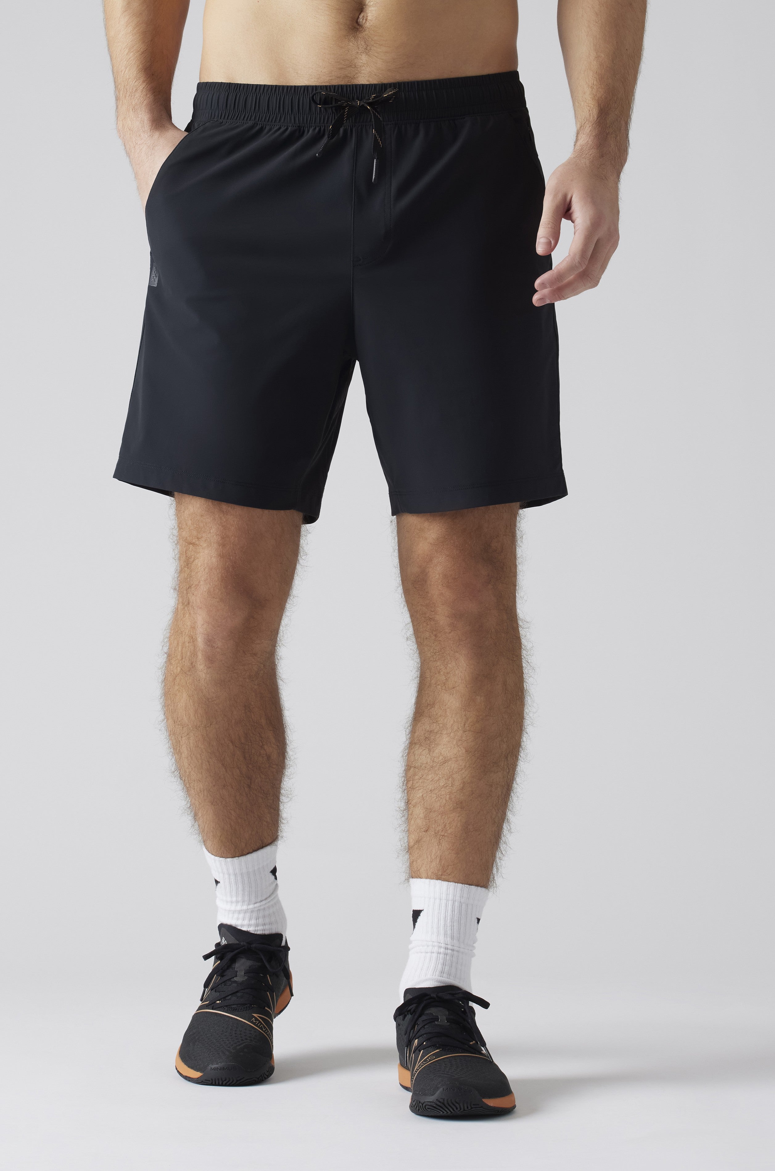 A man is seen from the waist down wearing South Moon Under Pursuit Shorts 7 In Unlined in black, white crew socks, and black athletic shoes, standing against a plain light gray background.