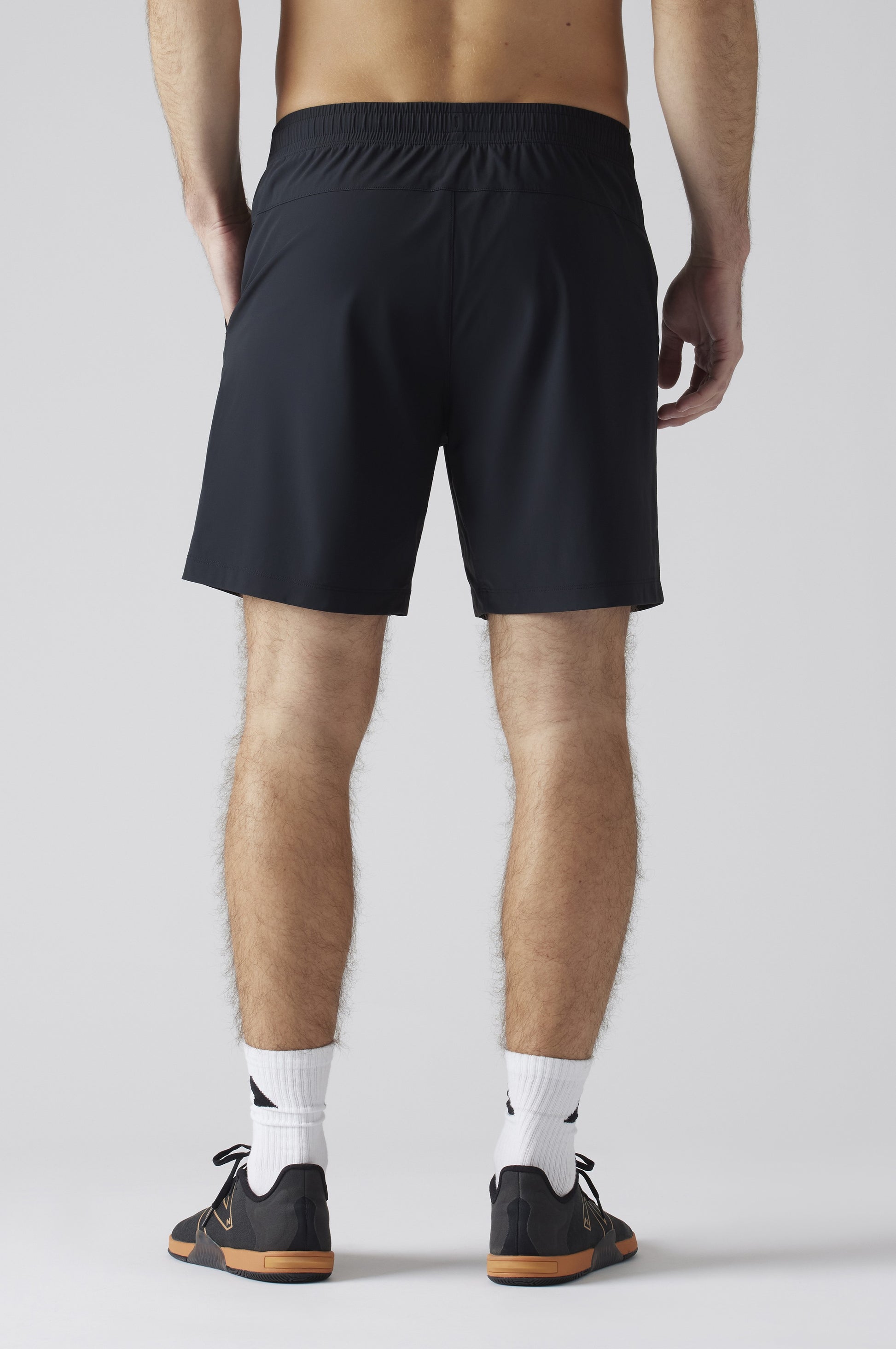 Someone stands facing away in South Moon Under Pursuit Shorts 7 In Unlined, black with an elastic waistband, paired with white crew socks and black athletic shoes with brown soles. The background is plain and light-colored.