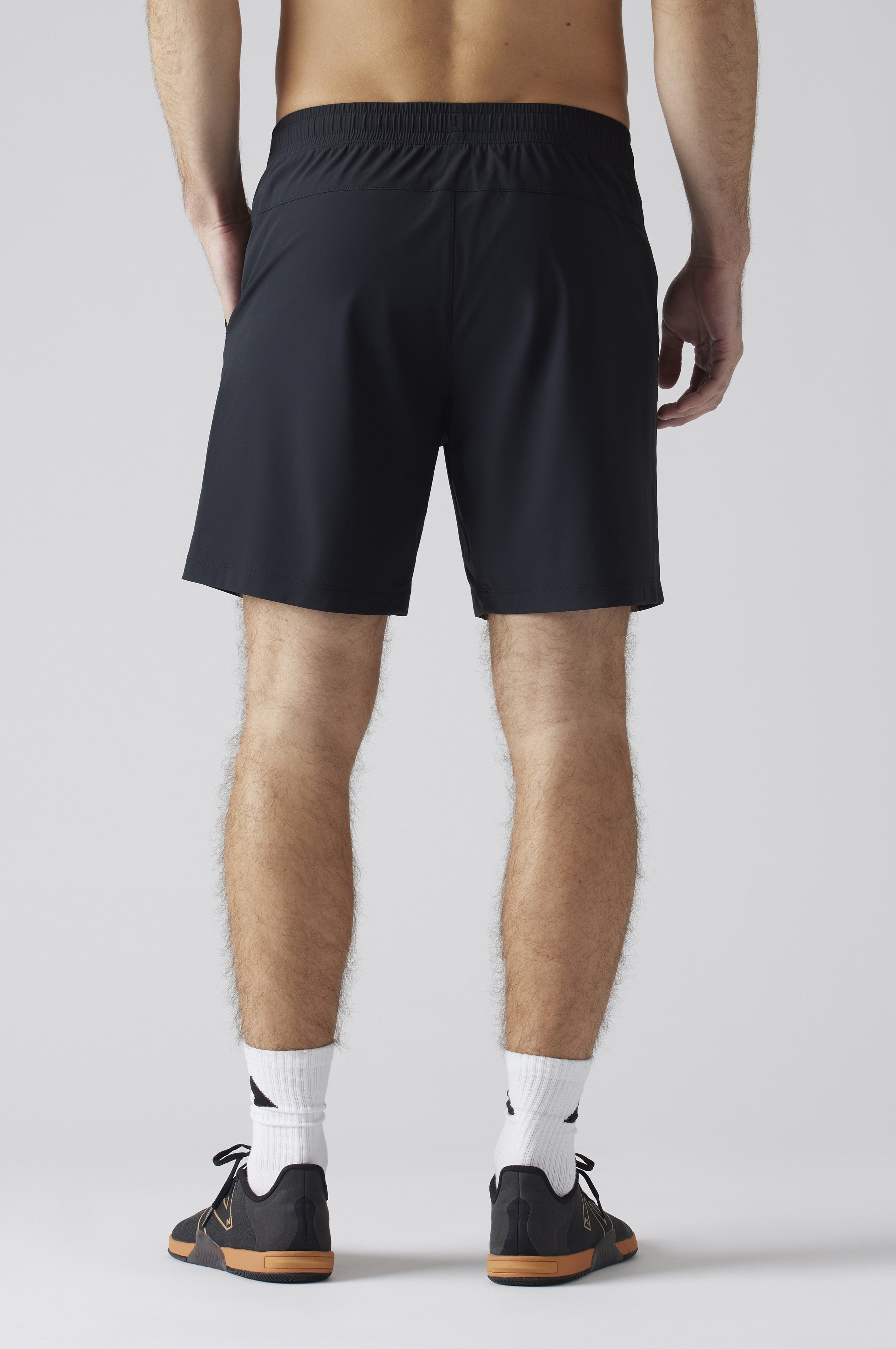 Someone stands facing away in South Moon Under Pursuit Shorts 7 In Unlined, black with an elastic waistband, paired with white crew socks and black athletic shoes with brown soles. The background is plain and light-colored.