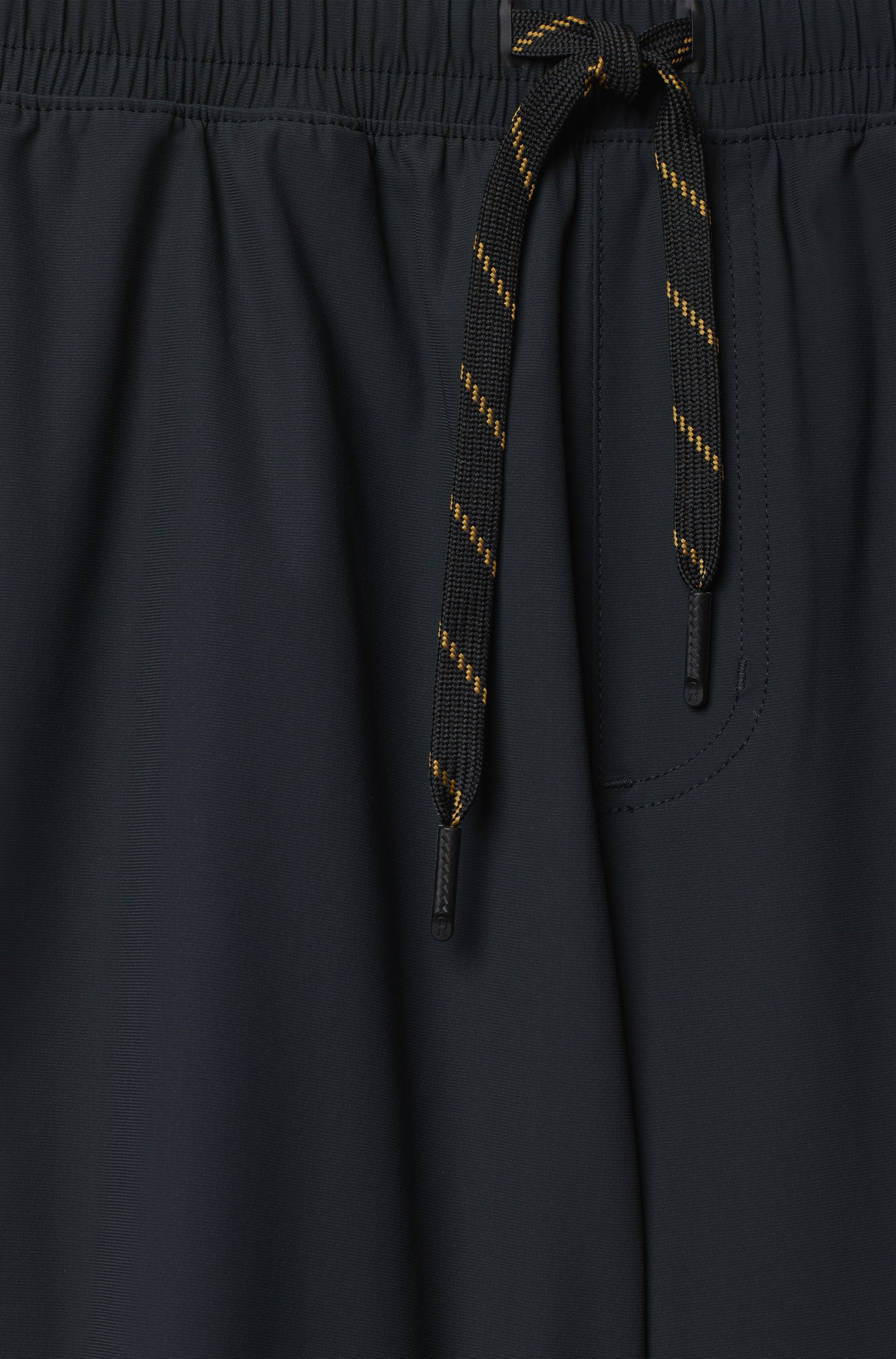 Close-up of South Moon Under Pursuit Shorts 7 In Unlined in black, featuring a standard fit, elastic waistband with black drawstring and yellow stripes, plus a zippered pocket with visible stitching.