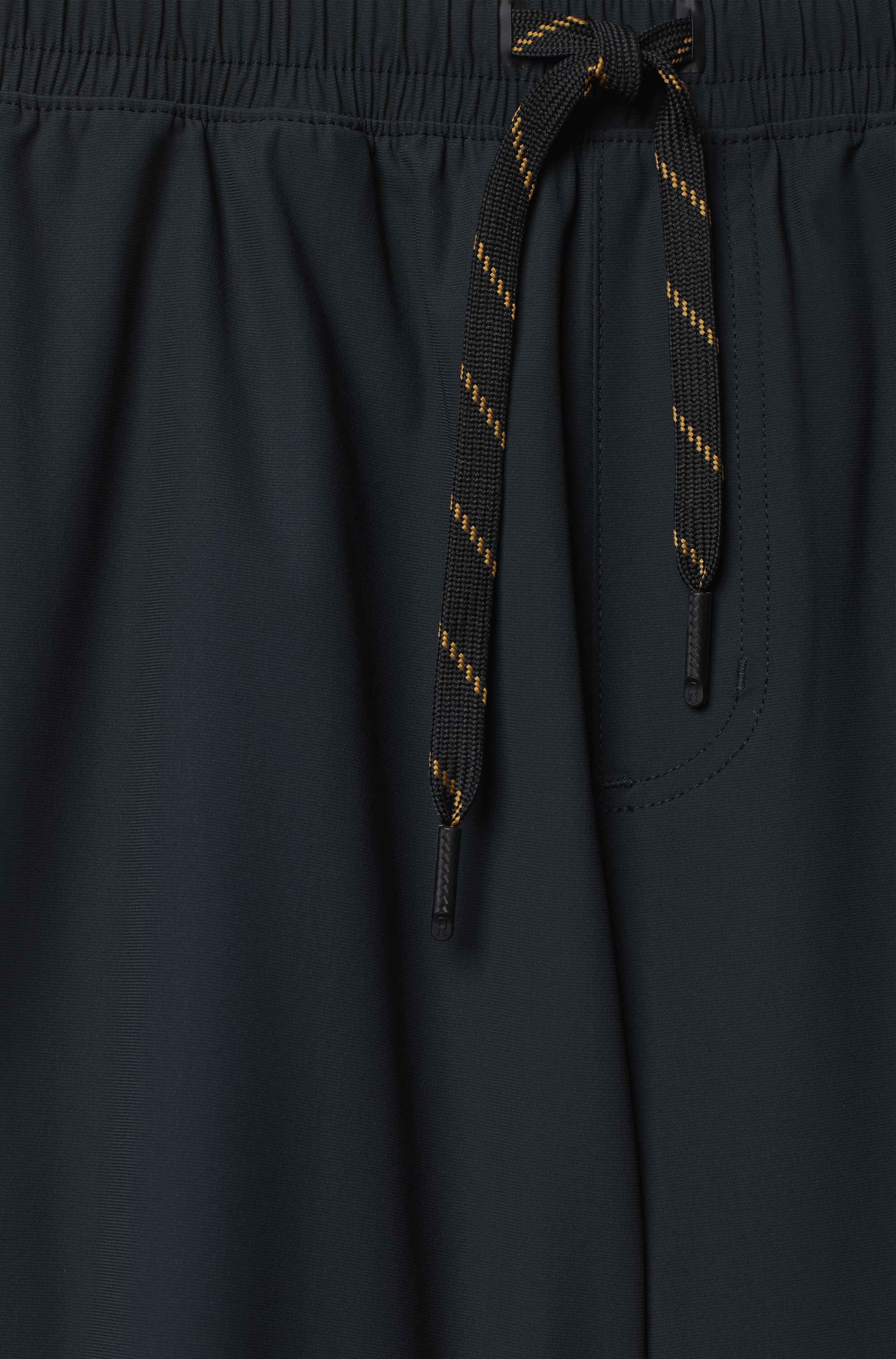 Close-up of South Moon Under Pursuit Shorts 7 In Unlined in black, featuring a standard fit, elastic waistband with black drawstring and yellow stripes, plus a zippered pocket with visible stitching.