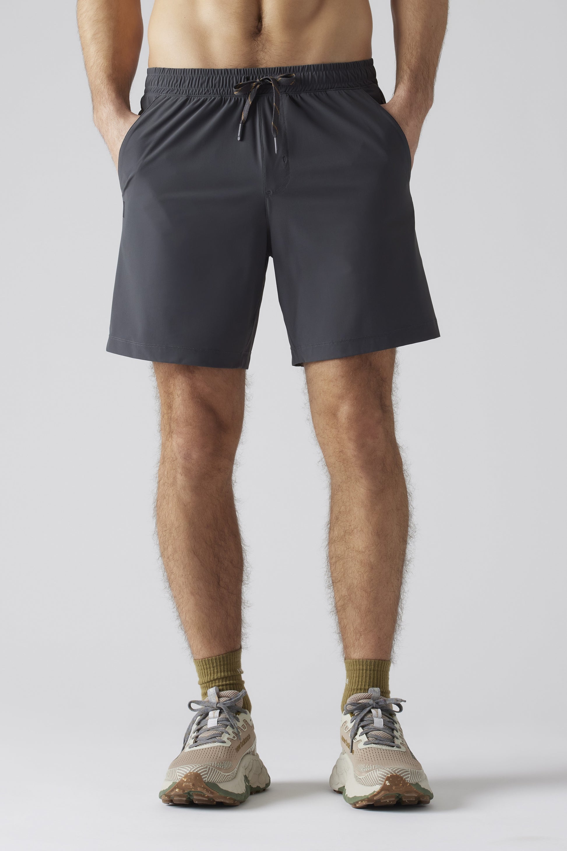 A person stands hands-in-pockets against a plain light gray background, wearing South Moon Under Pursuit Shorts 7 In Unlined in navy, olive green socks, and beige running shoes. The image is cropped from shoulders to feet.
