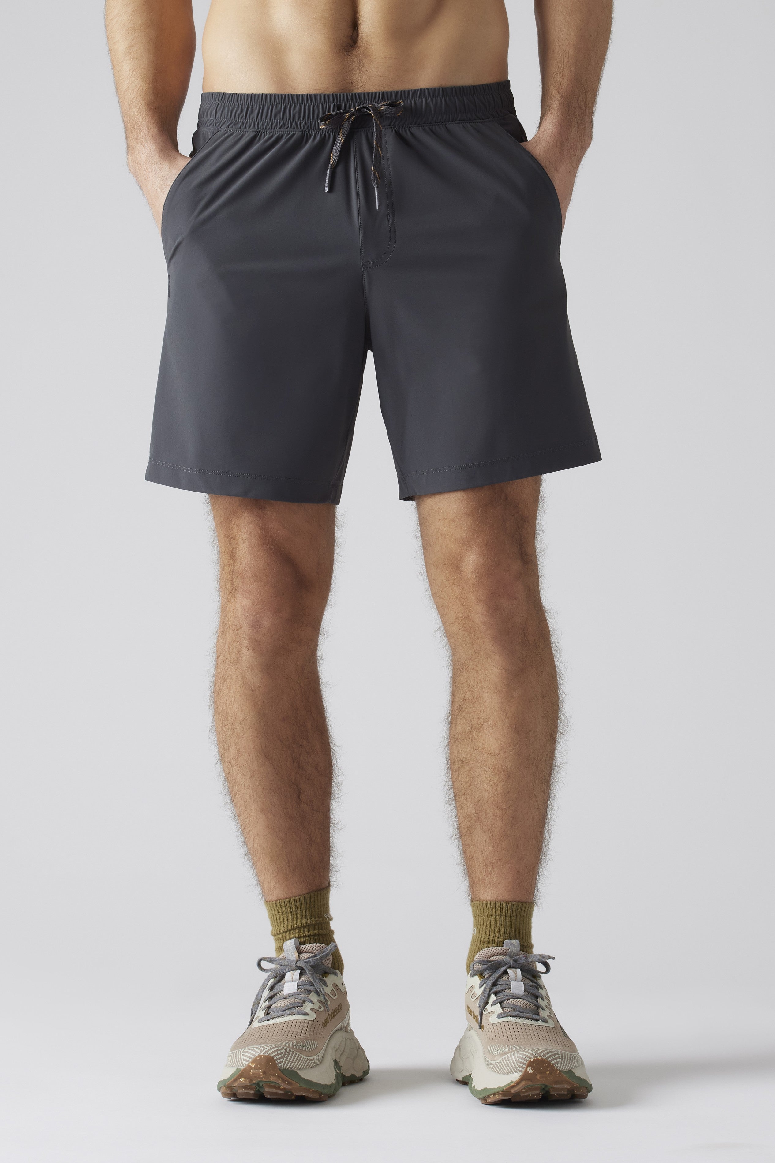A person stands hands-in-pockets against a plain light gray background, wearing South Moon Under Pursuit Shorts 7 In Unlined in navy, olive green socks, and beige running shoes. The image is cropped from shoulders to feet.