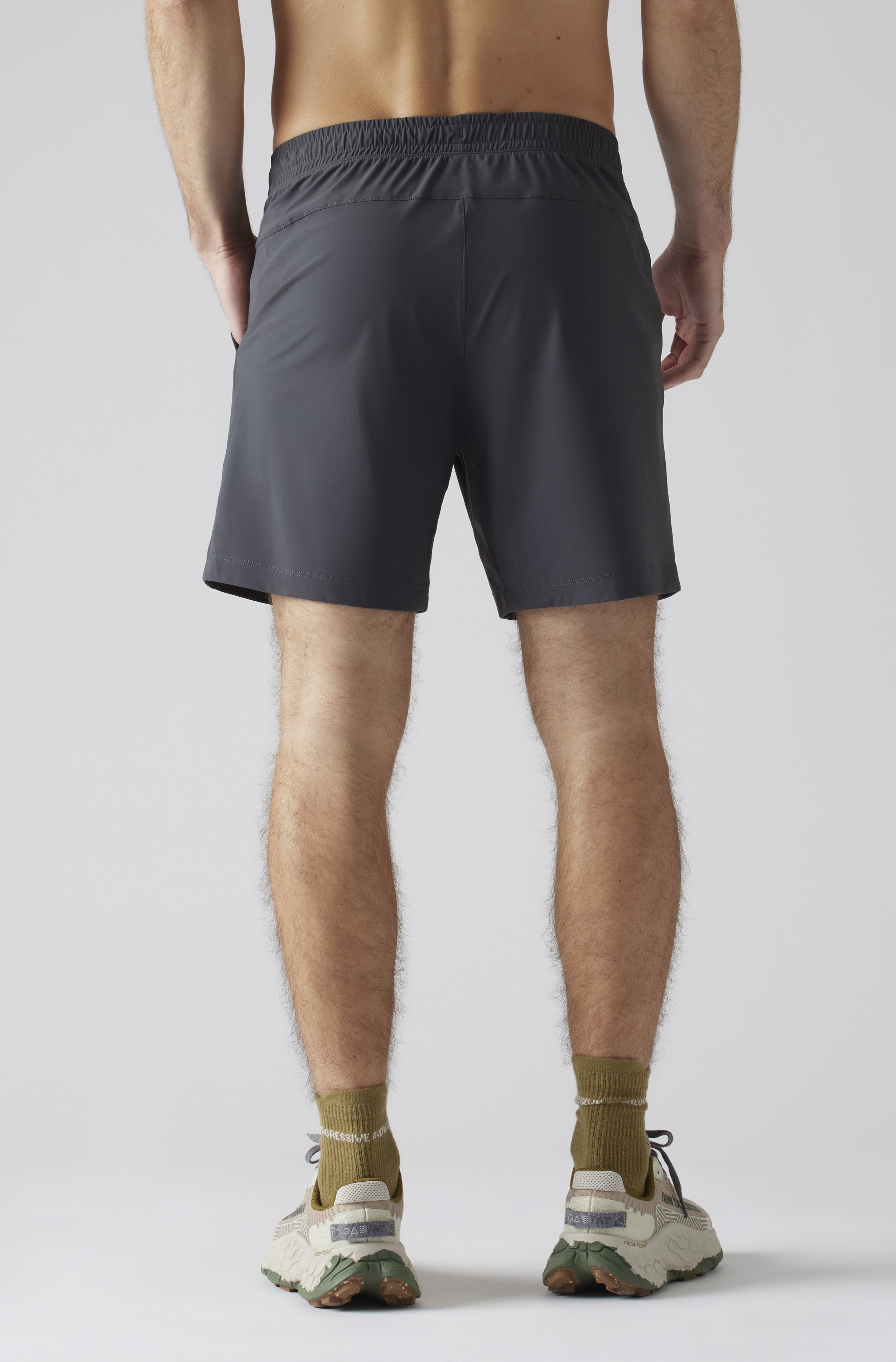 A person faces away, wearing South Moon Under Pursuit Shorts 7 In Unlined in dark gray with a zipper pocket, paired with olive socks and light running shoes, against a plain light background.