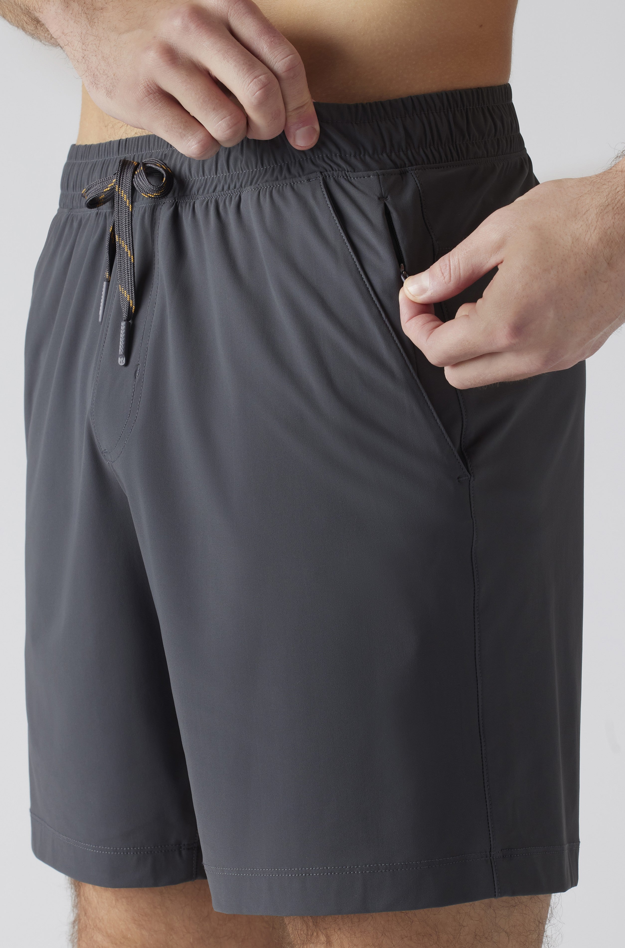 A person is shown from waist to mid-thigh wearing South Moon Under Pursuit Shorts 7 In Unlined in dark gray, adjusting the elastic waistband with one hand while placing fingers in a zipper pocket with the other.