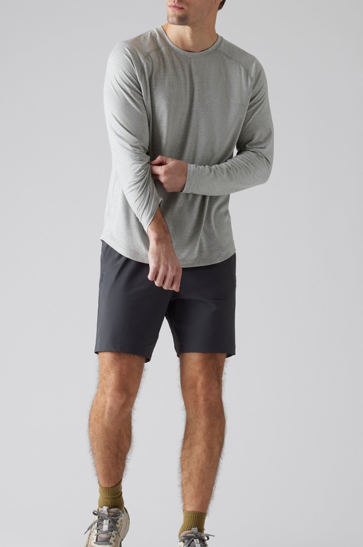 A man in light gray long-sleeve shirt and South Moon Under Pursuit Shorts 7 In Unlined with a zipper pocket stands against a plain light background, adjusting his sleeve. His head is slightly turned, and his face isn't fully visible.