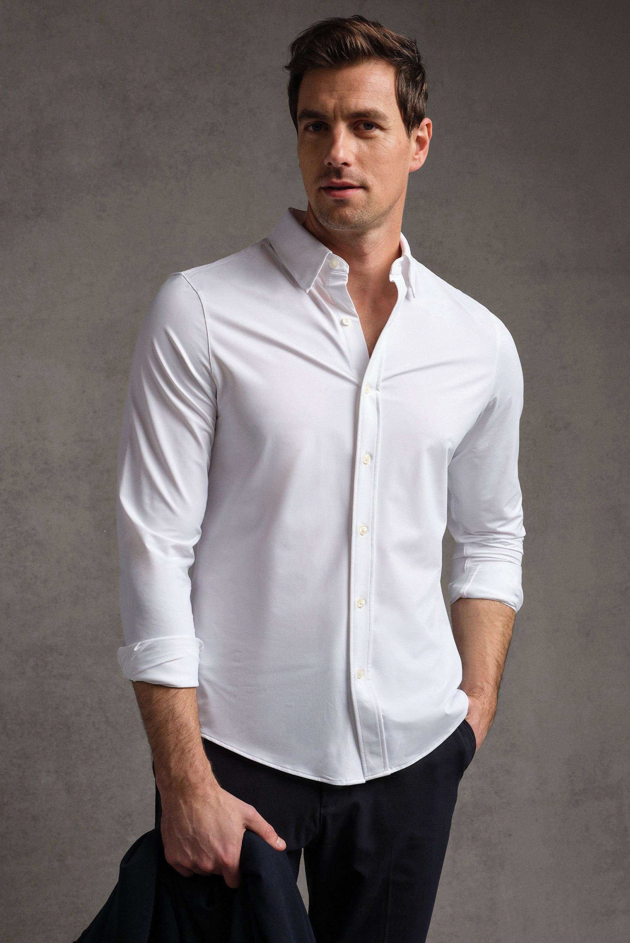 A man wearing the RHONE Commuter Shirt- Classic Fit in white with rolled-up sleeves stands against a plain gray background, confidently holding a dark jacket and looking at the camera.