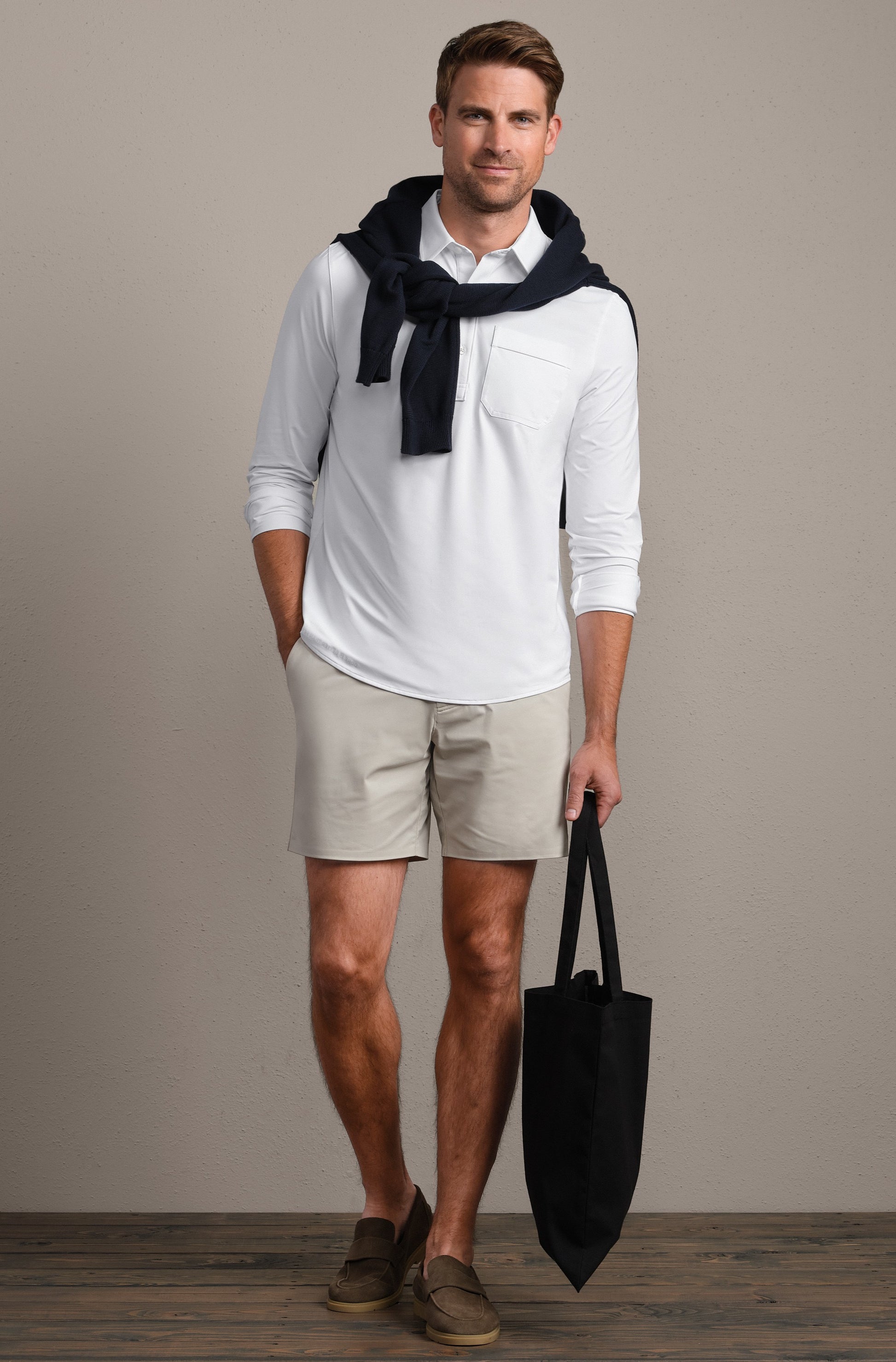 A man stands indoors wearing the RHONE Commuter Shirt- Classic Fit in white, paired with light beige shorts, brown loafers, a navy sweater draped over his shoulders, and holding a black tote bag in his left hand.