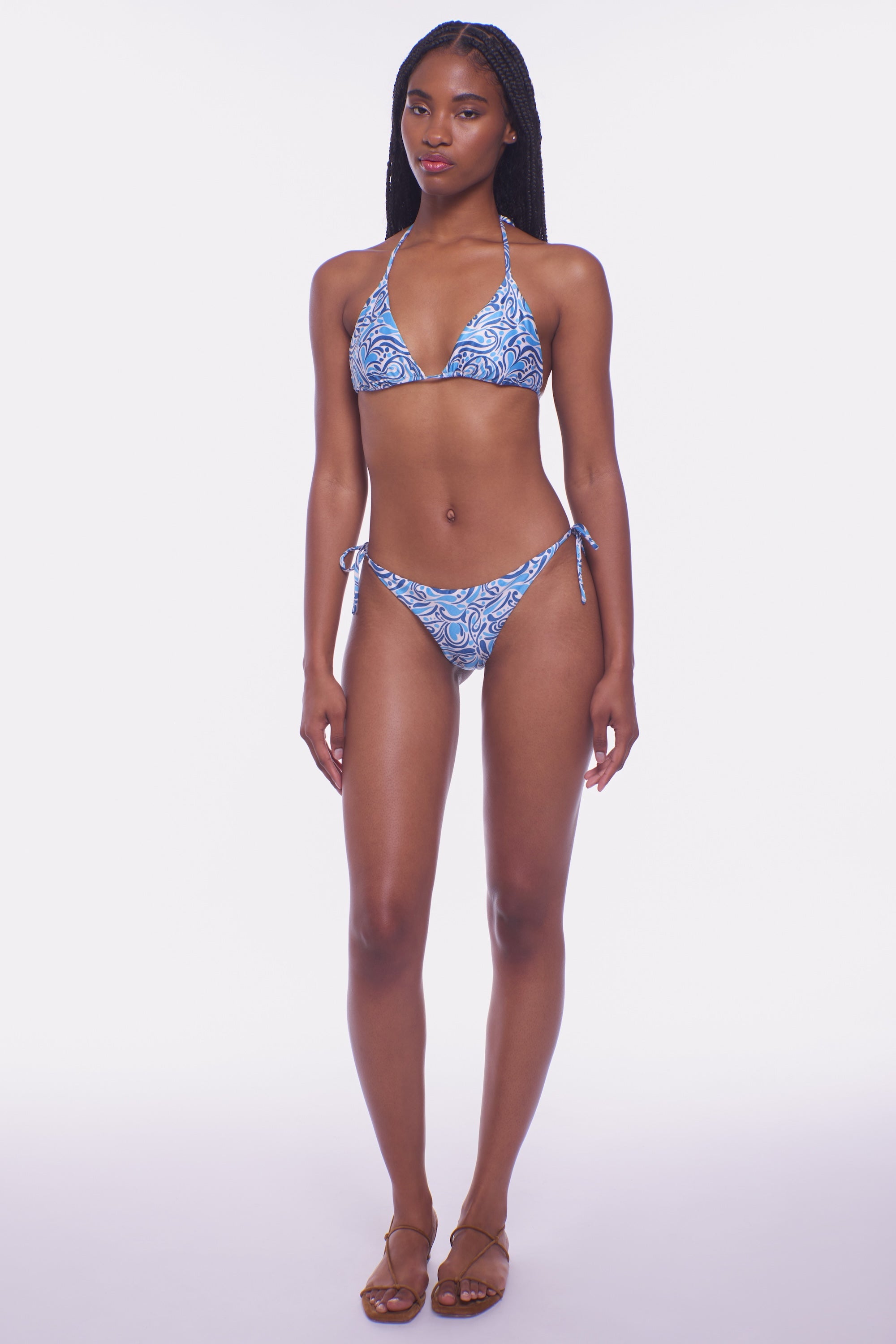 A woman wears the Peixoto Fifi Bikini Top, featuring a blue and white pattern, with brown sandals against a white background. This polyester bikini top is best cared for by gentle hand washing.
