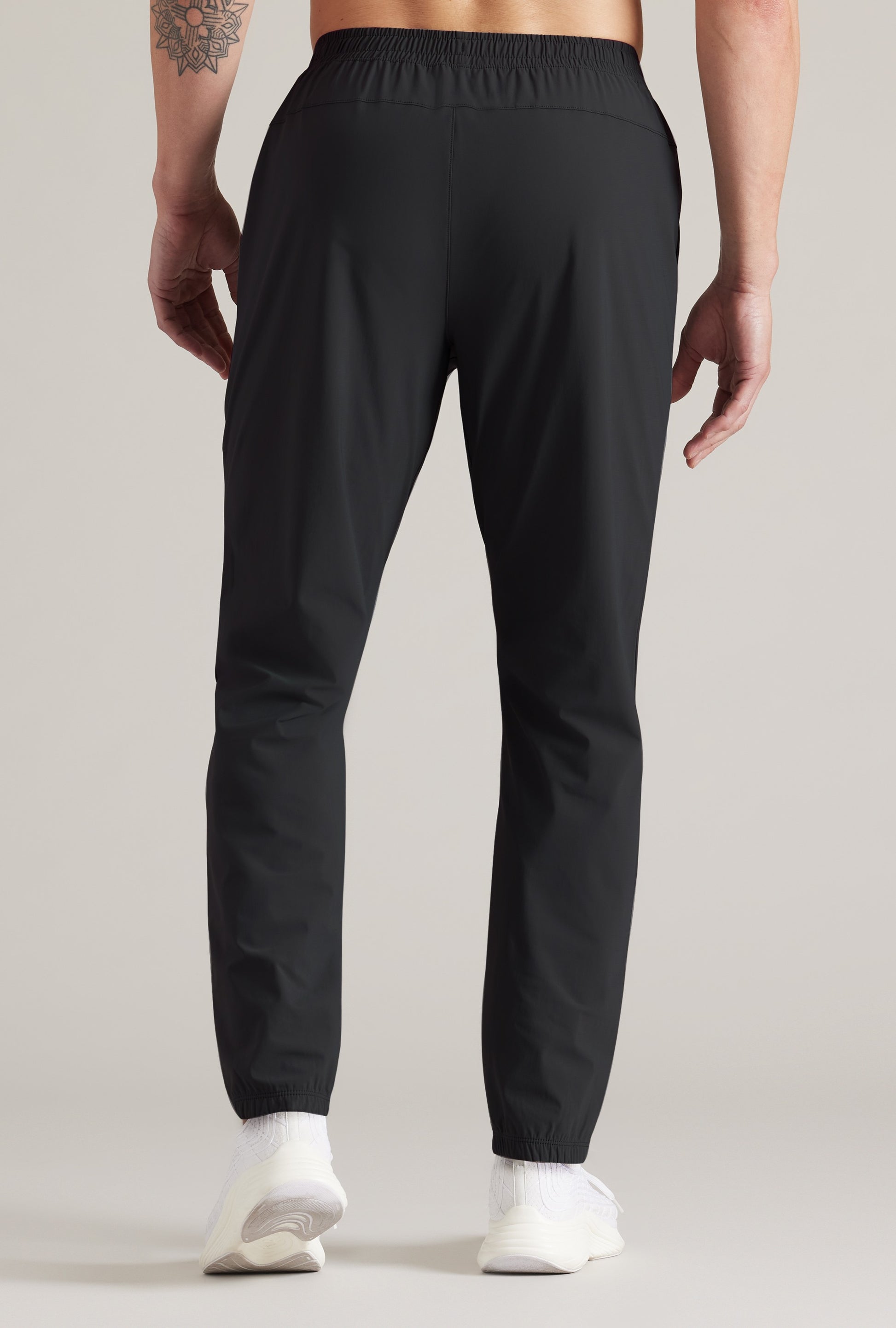 Someone stands facing away from the camera in South Moon Under Pursuit Pant, showcasing the back view. The black athletic-fit joggers are paired with white socks; a tattoo is visible on their left arm. Background is plain and light-colored.