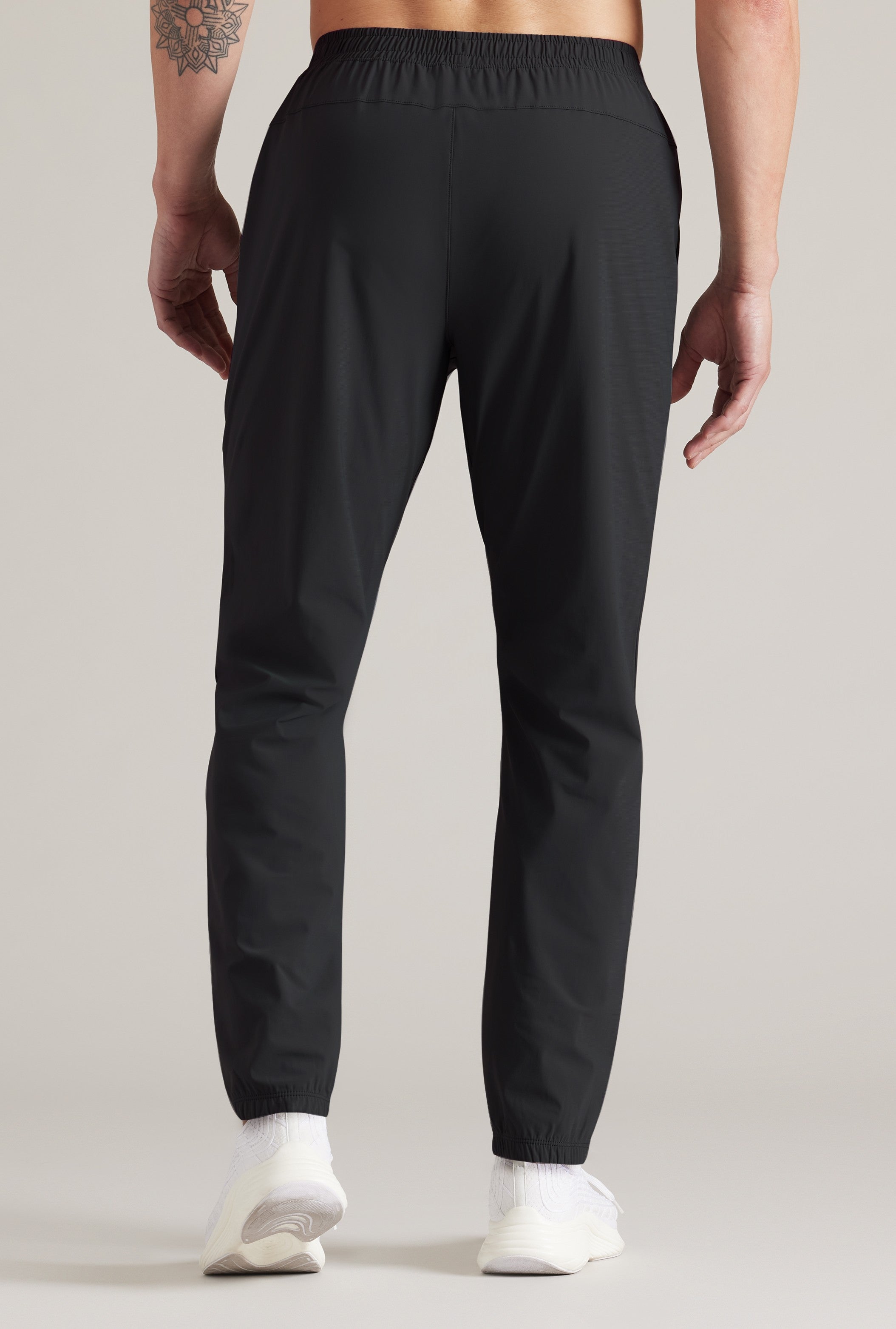 Someone stands facing away from the camera in South Moon Under Pursuit Pant, showcasing the back view. The black athletic-fit joggers are paired with white socks; a tattoo is visible on their left arm. Background is plain and light-colored.