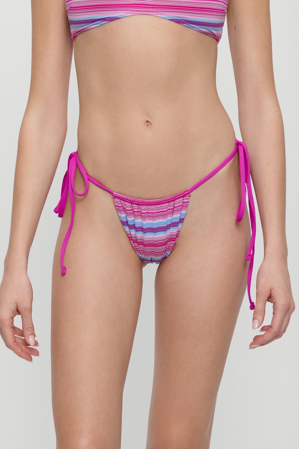Model wears the Frankies Bikinis Tia Space Dyed Bottom, a colorful striped ruched bikini bottom with pink string side ties, shown from shoulders to upper thighs against a plain background.
