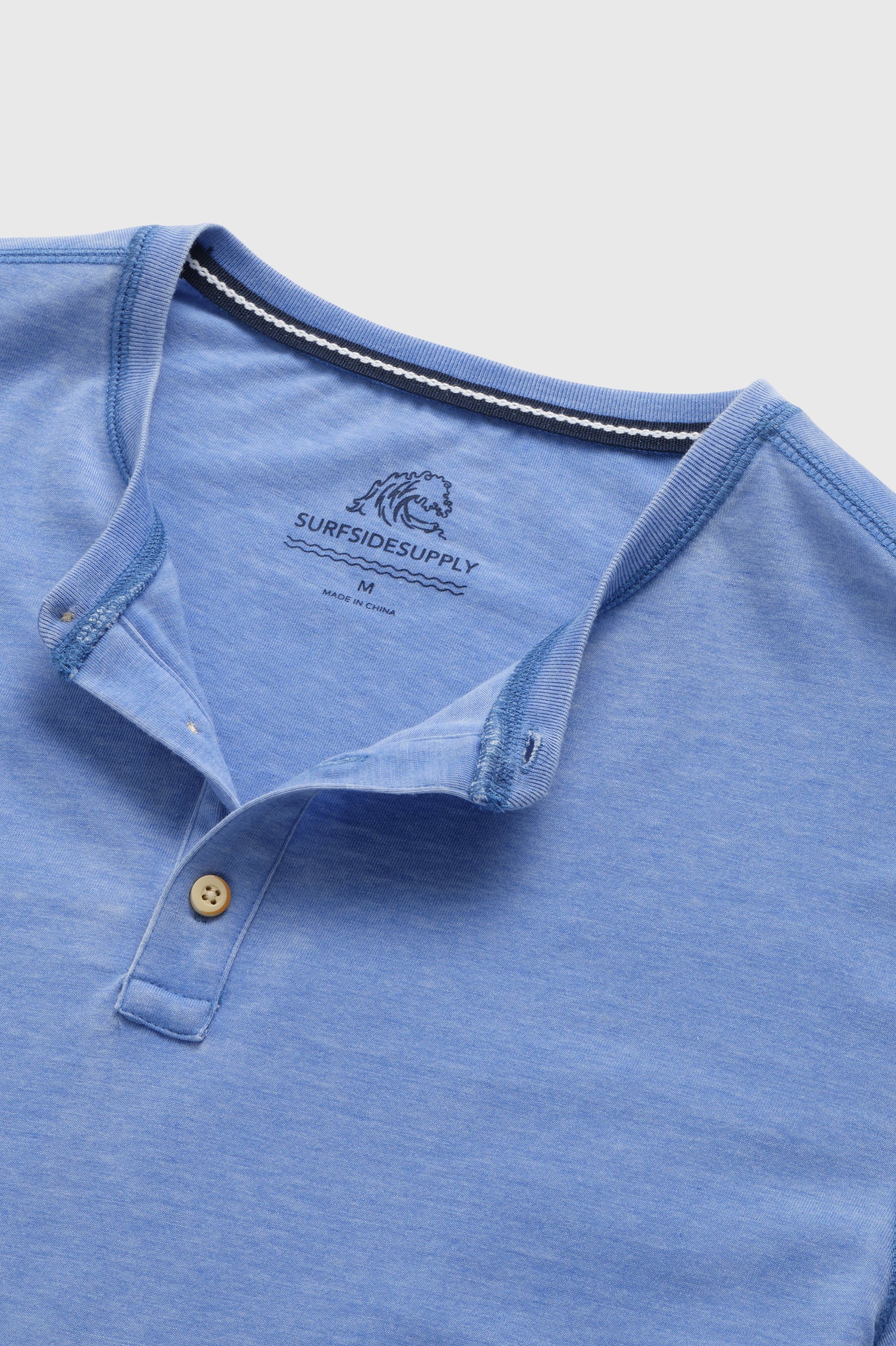 A close-up of the Sean Short Sleeve Burnout Henley by Surfside Supply Co., a light blue knit with short sleeves, two-button placket, and a “SURFSIDE SUPPLY” logo plus wave graphic printed inside the collar.