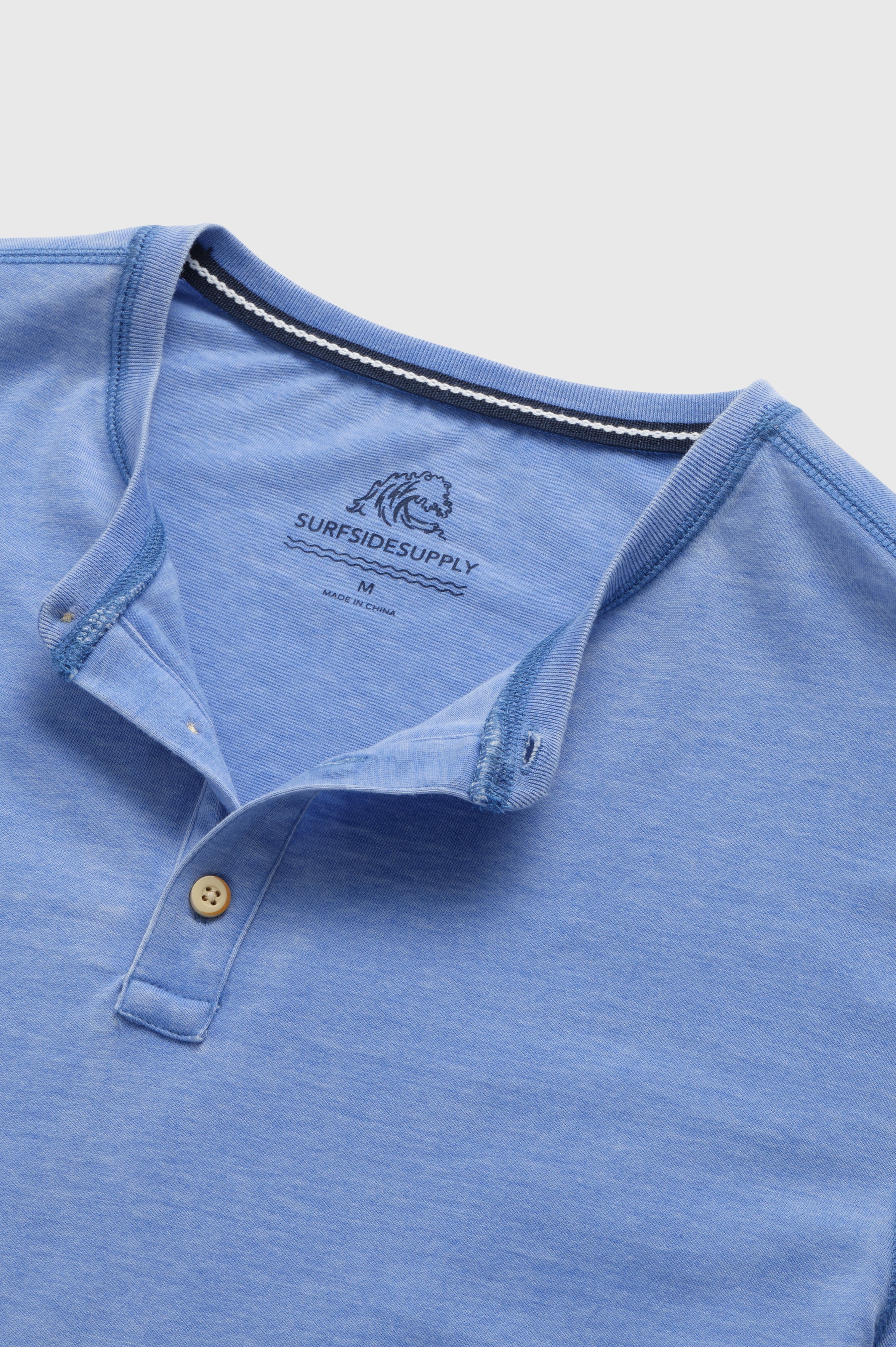 A close-up of the Sean Short Sleeve Burnout Henley by Surfside Supply Co., a light blue knit with short sleeves, two-button placket, and a “SURFSIDE SUPPLY” logo plus wave graphic printed inside the collar.