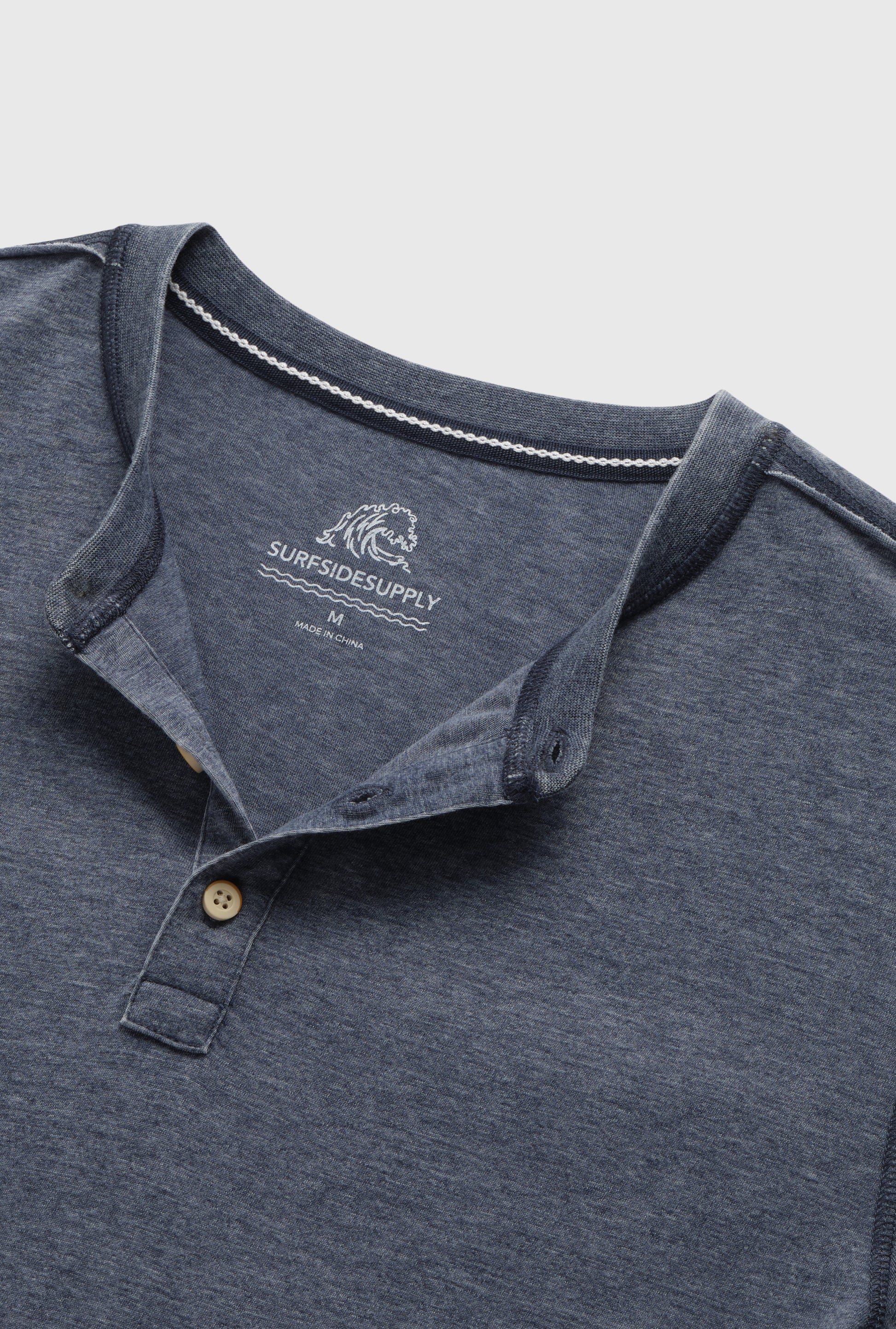 A close-up of the Surfside Supply Co. Sean Short Sleeve Burnout Henley in blue, featuring a partially open collar, two beige buttons, and inner neckline branding. The shirt is laid flat on a light background.