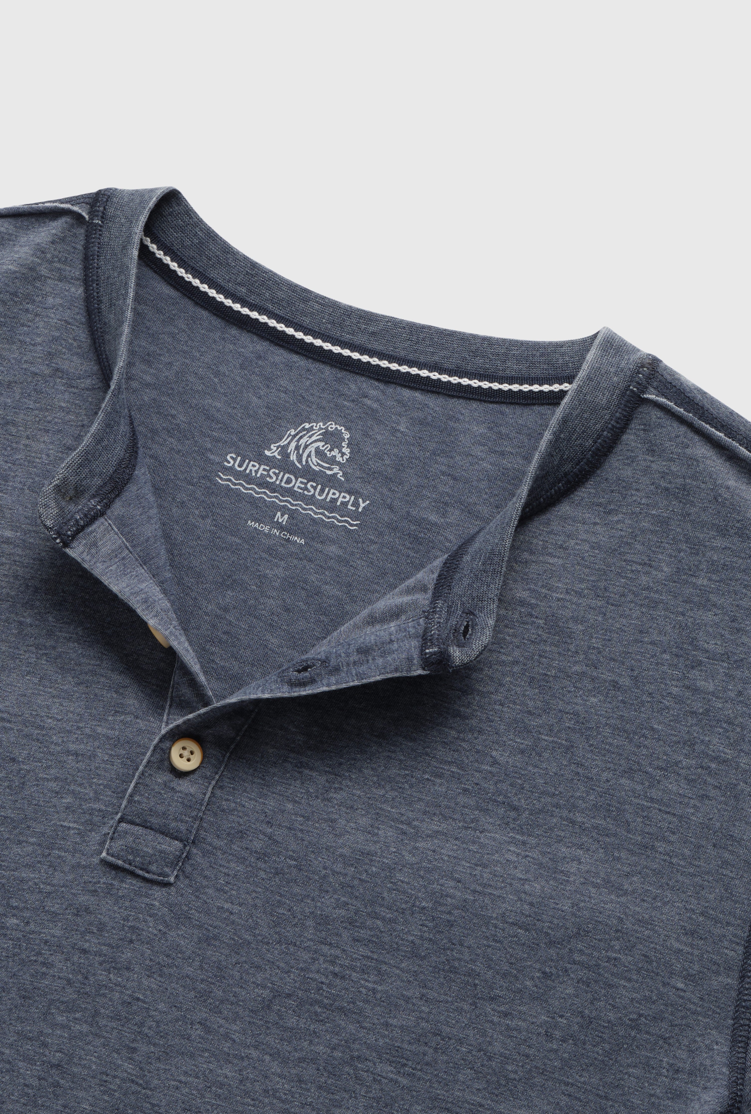 A close-up of the Surfside Supply Co. Sean Short Sleeve Burnout Henley in blue, featuring a partially open collar, two beige buttons, and inner neckline branding. The shirt is laid flat on a light background.