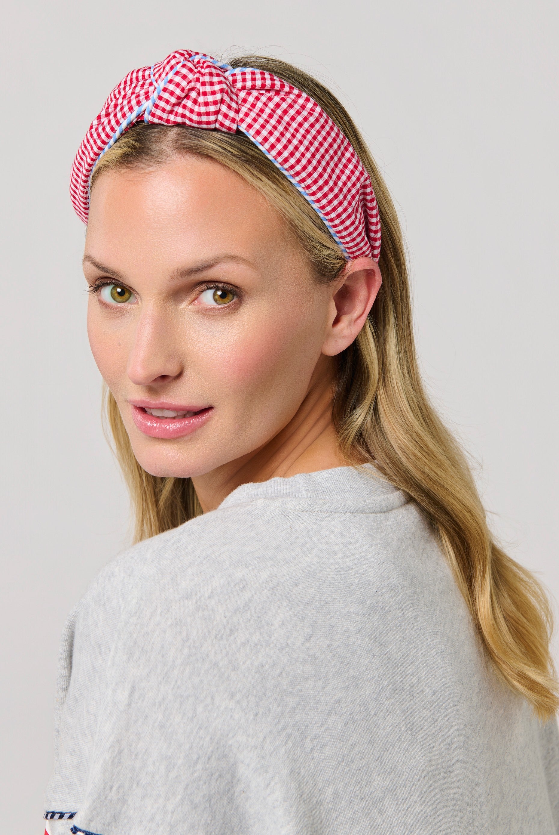 A woman with long blonde hair wears the South Moon Under Gingham Knotted Headband and a light gray top, glancing over her shoulder at the camera—the perfect summer accessory against a plain, light background.
