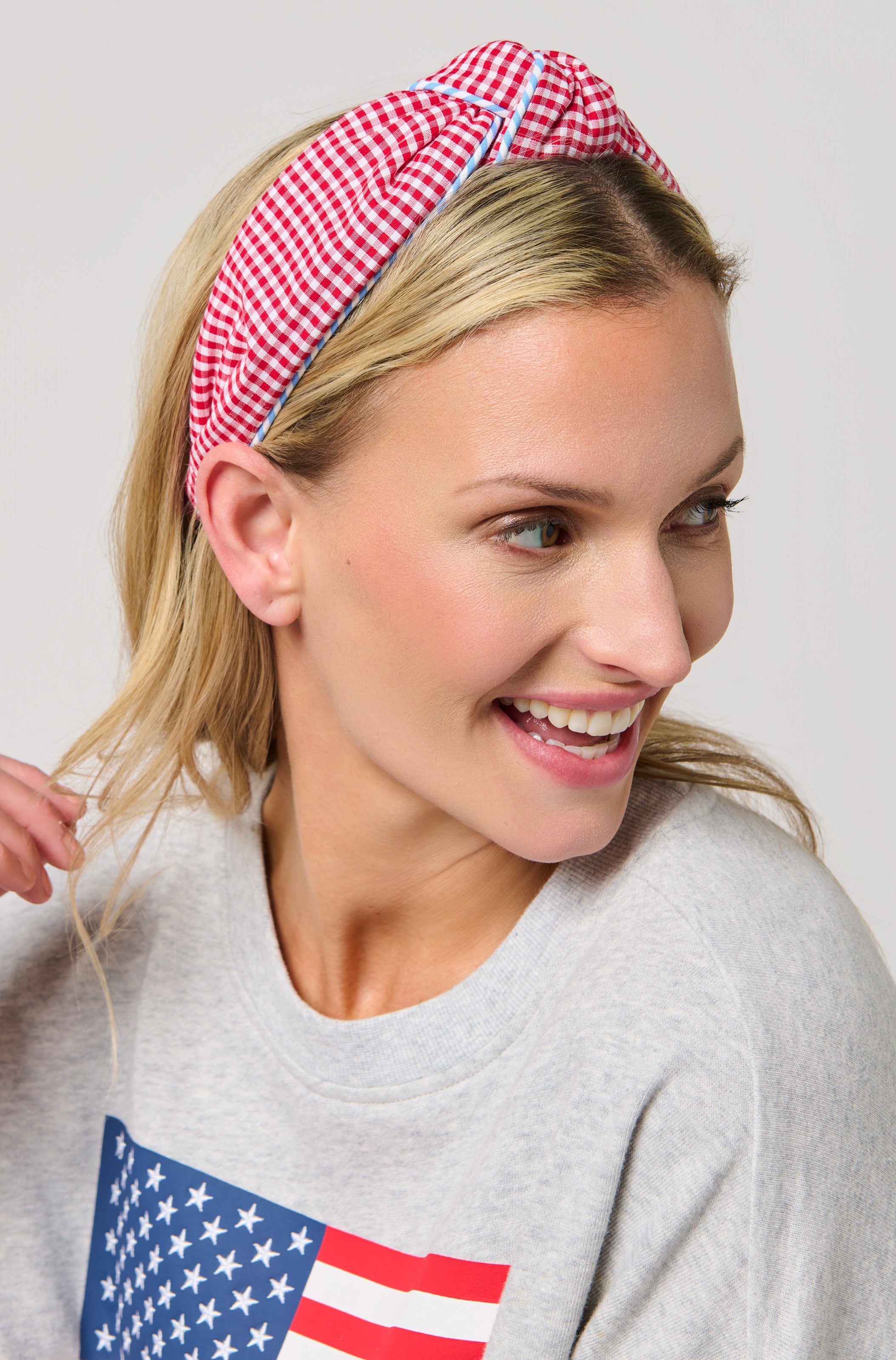 A smiling woman with blonde hair wears the South Moon Under Gingham Knotted Headband in Red Multi, paired with a light gray sweatshirt with an American flag design. She looks to the side, radiating joy—the perfect fun summer accessory.