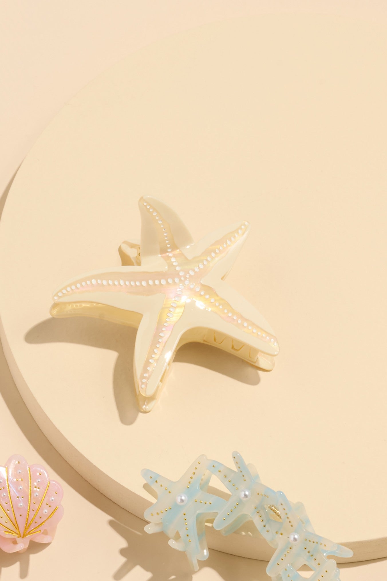 The Shiraleah Starfish Claw Clip in ivory and two other pastel ocean-themed hair accessories are displayed on a round, cream-colored surface.