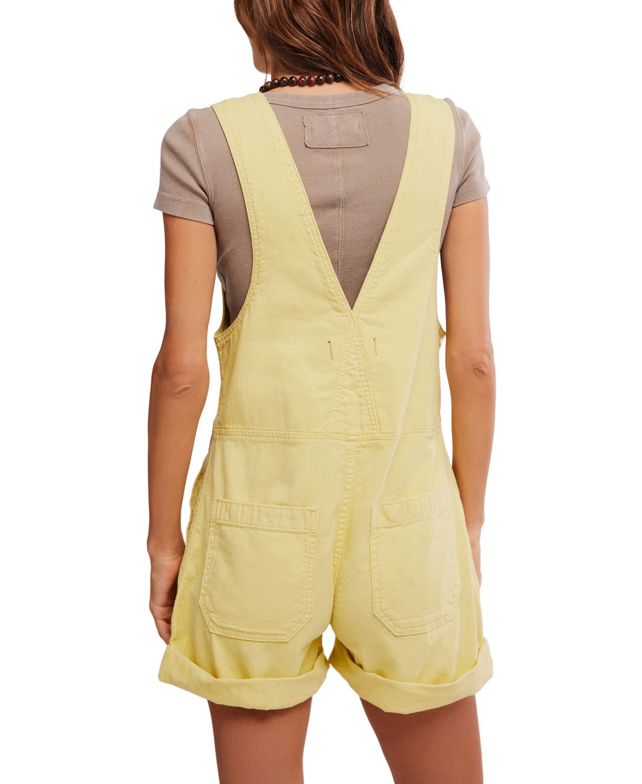 A person is shown from the back wearing the Jude Shortall by Free People: light yellow denim overalls with rolled cuffs and two back pockets, styled over a short-sleeve taupe shirt, against a plain white background.