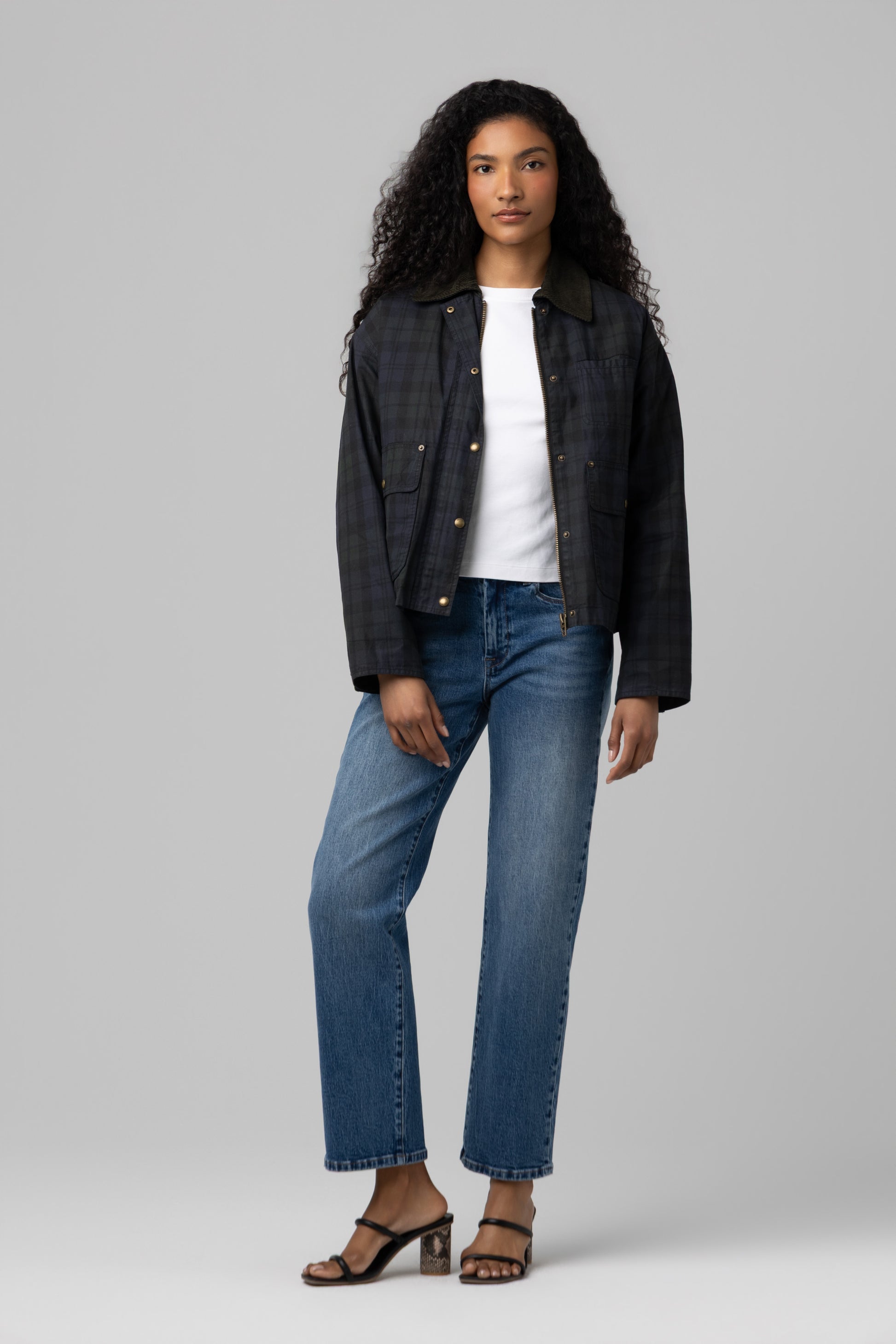 A person stands facing forward against a plain gray background, wearing the Break Trail Barn Jacket by Blank—a dark plaid piece with a cotton shell—layered over a white shirt, blue jeans, and heeled sandals. They have long curly hair and a neutral expression.