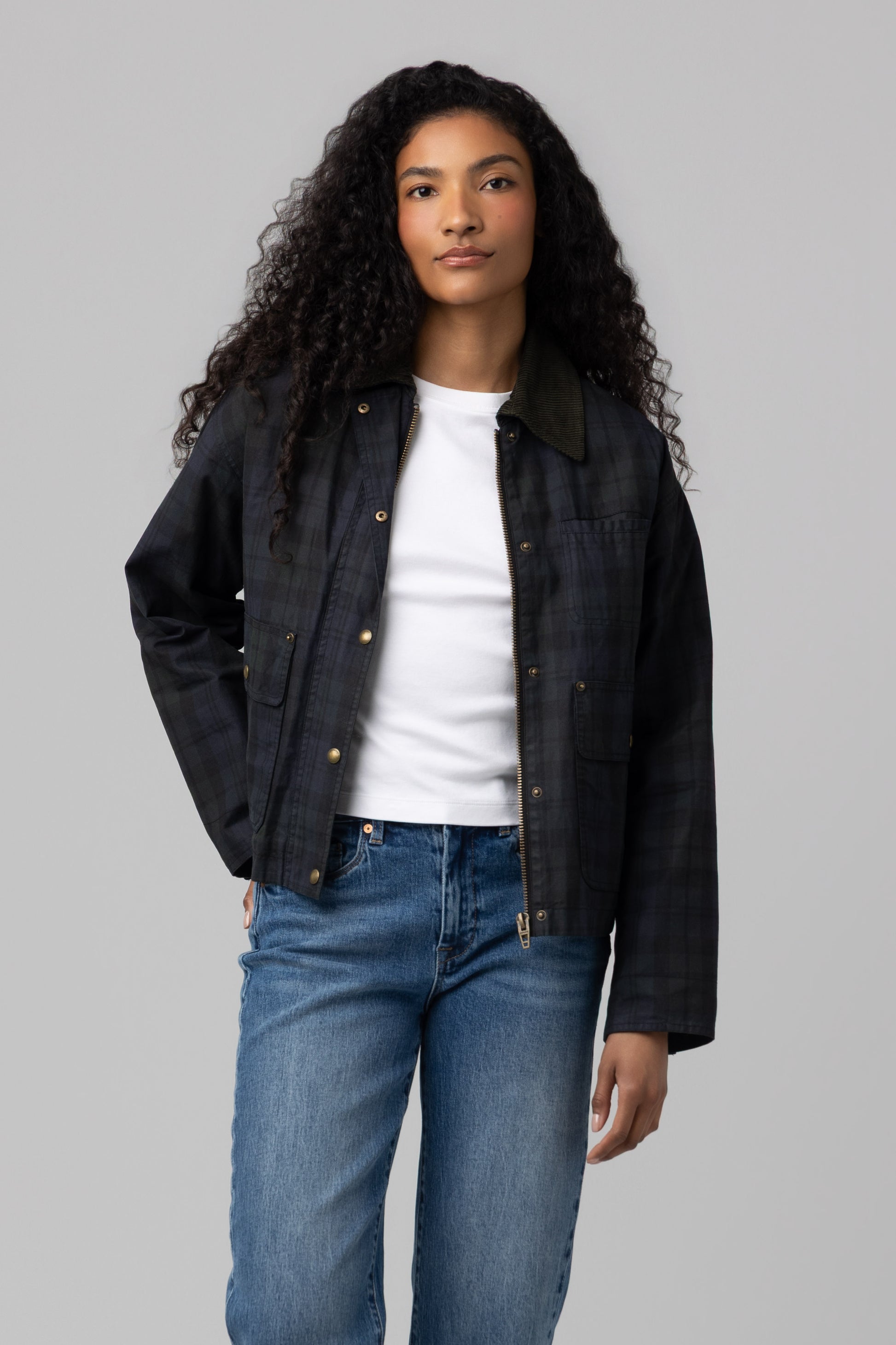 A person with long curly hair wears the BREAK TRAIL-colored Break Trail Barn Jacket by Blank, featuring a soft cotton shell, layered over a white t-shirt and blue jeans while standing against a plain gray background.