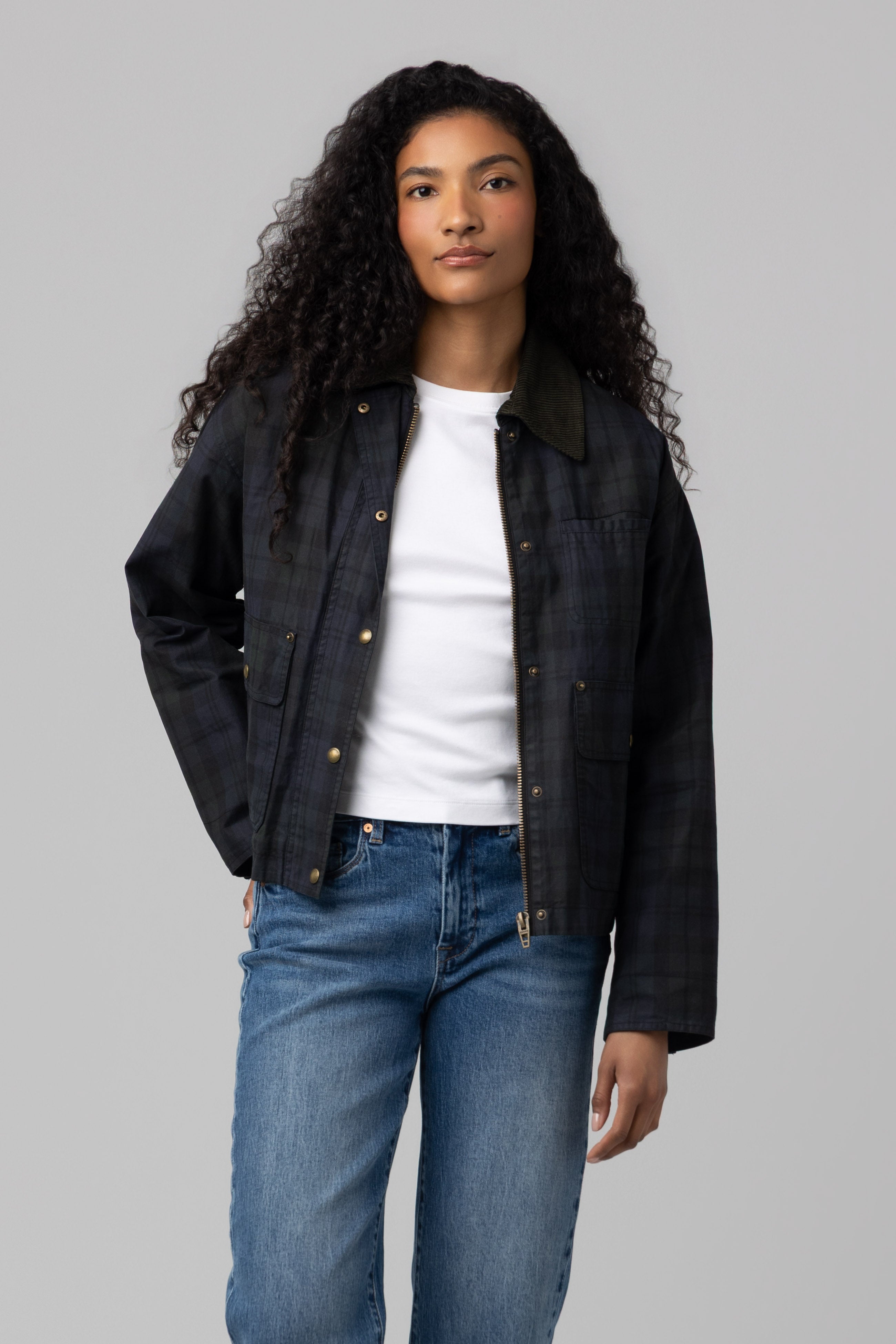 A person with long curly hair wears the BREAK TRAIL-colored Break Trail Barn Jacket by Blank, featuring a soft cotton shell, layered over a white t-shirt and blue jeans while standing against a plain gray background.