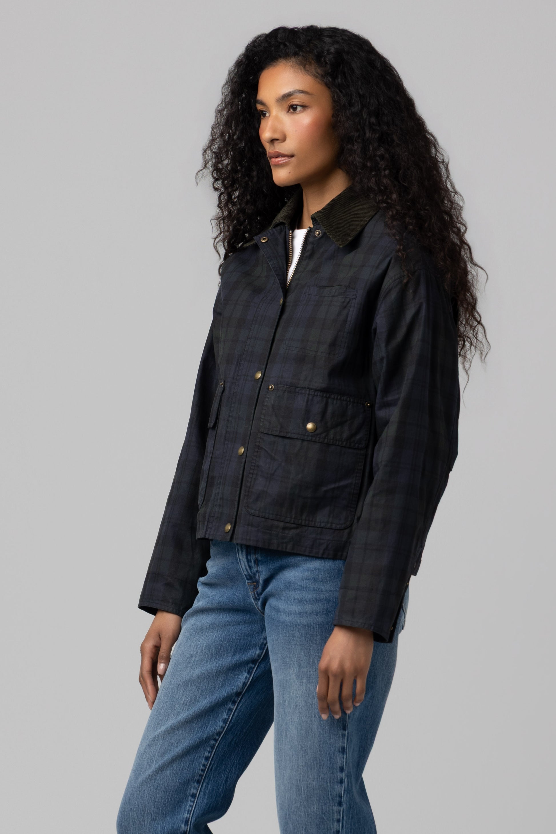 A woman with long curly hair stands against a plain light gray background, wearing the Blank Break Trail Barn Jacket—a dark plaid style featuring front pockets and blue jeans. The jacket's cotton shell and insulated construction provide both warmth and style.
