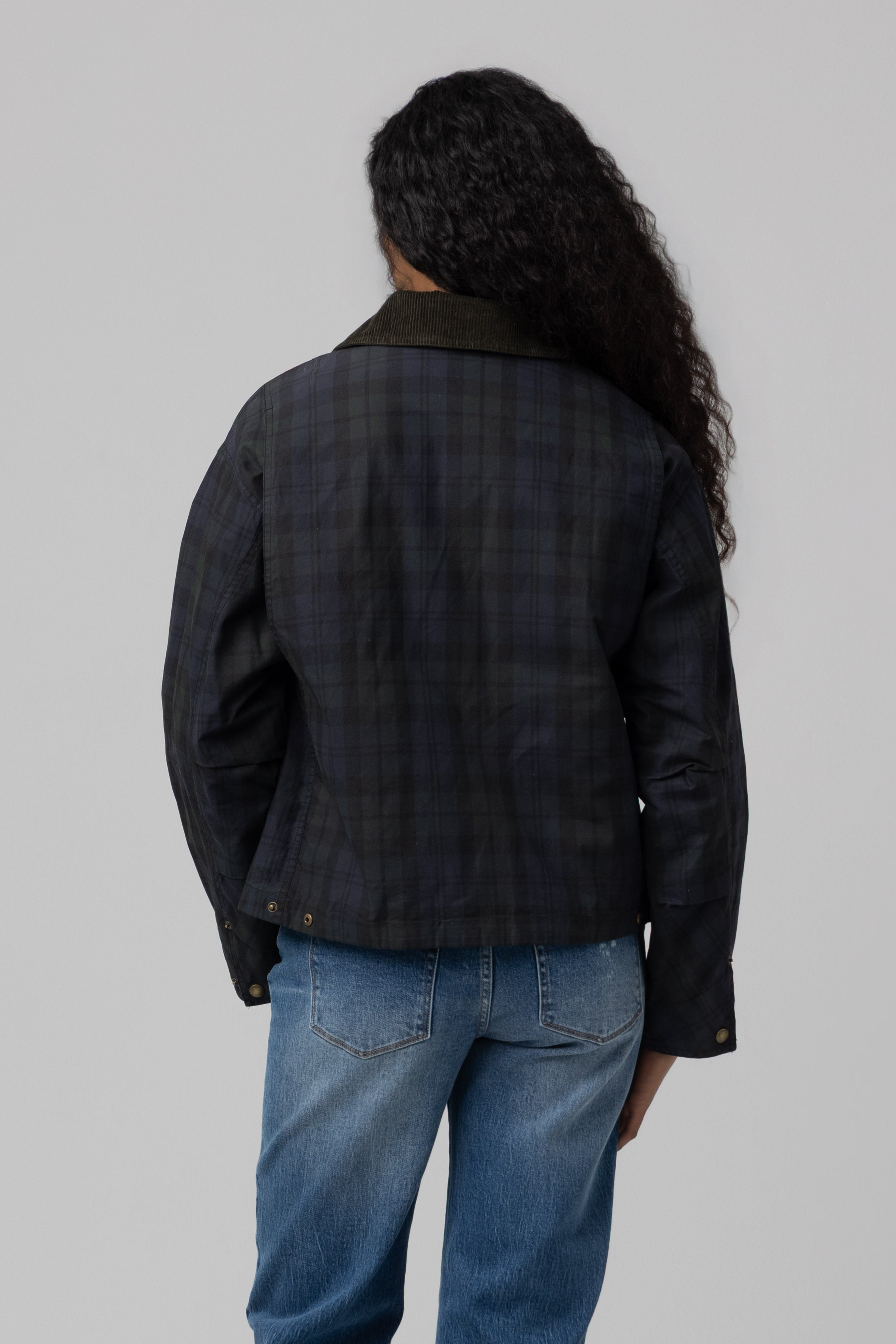 A person with long curly hair, dressed in the Blank Break Trail Barn Jacket featuring a durable cotton shell in dark plaid and blue jeans, stands facing away from the camera against a plain gray background.