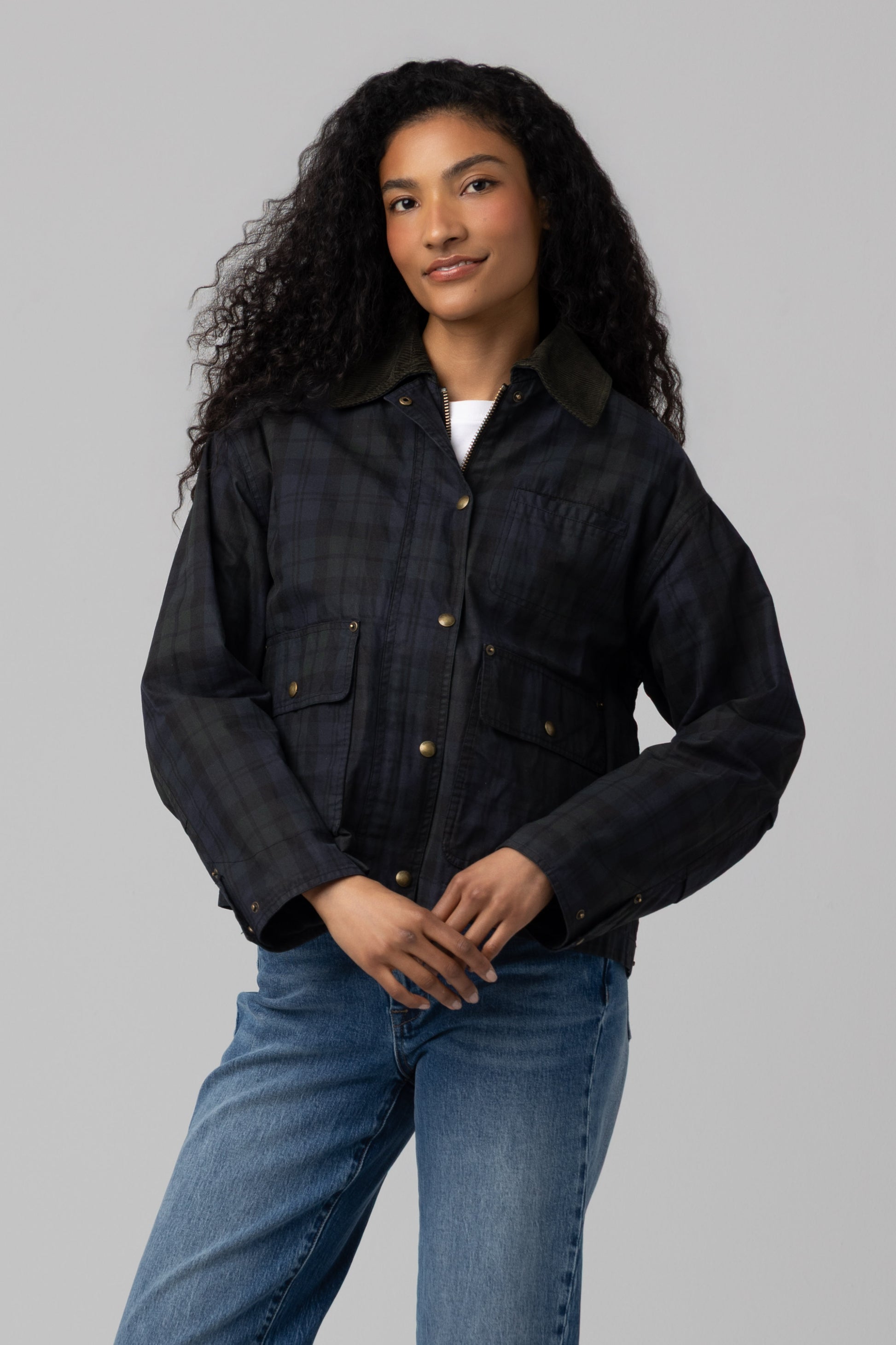 A person with long curly hair wears the Blank Break Trail Barn Jacket—a dark plaid style featuring an insulated construction and cotton shell—layered over a white shirt and blue jeans, standing against a plain gray background with a relaxed expression.