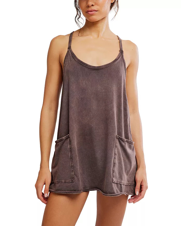 A woman stands against a plain white background wearing the FP Movement Hot Shot Mini, a loose, sleeveless brown cotton elastane tunic dress with thin straps, front pockets, and a relaxed silhouette.