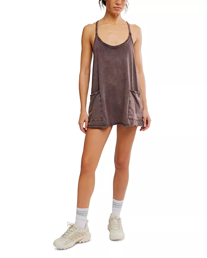 Someone is wearing the FP Movement Hot Shot Mini, a loose brown mini dress with front pockets and a relaxed fit, paired with white socks and beige sneakers, standing against a plain white background.