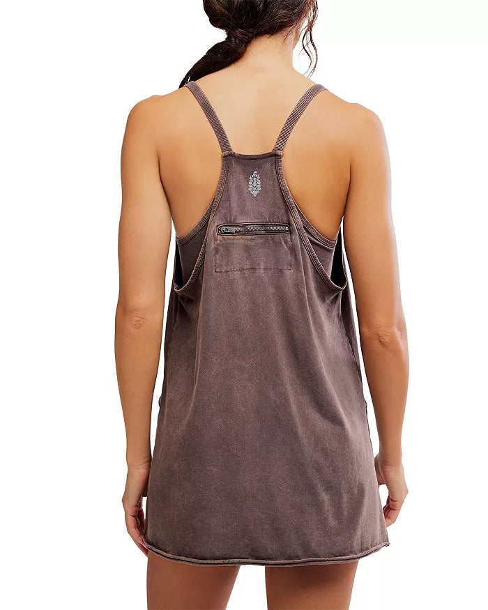 A woman, hair loosely tied, is seen from the back wearing the FP Movement Hot Shot Mini—a sleeveless faded brown cotton-elastane mini dress with racerback straps and a small zippered pocket on the upper back. The hem falls above her knees.