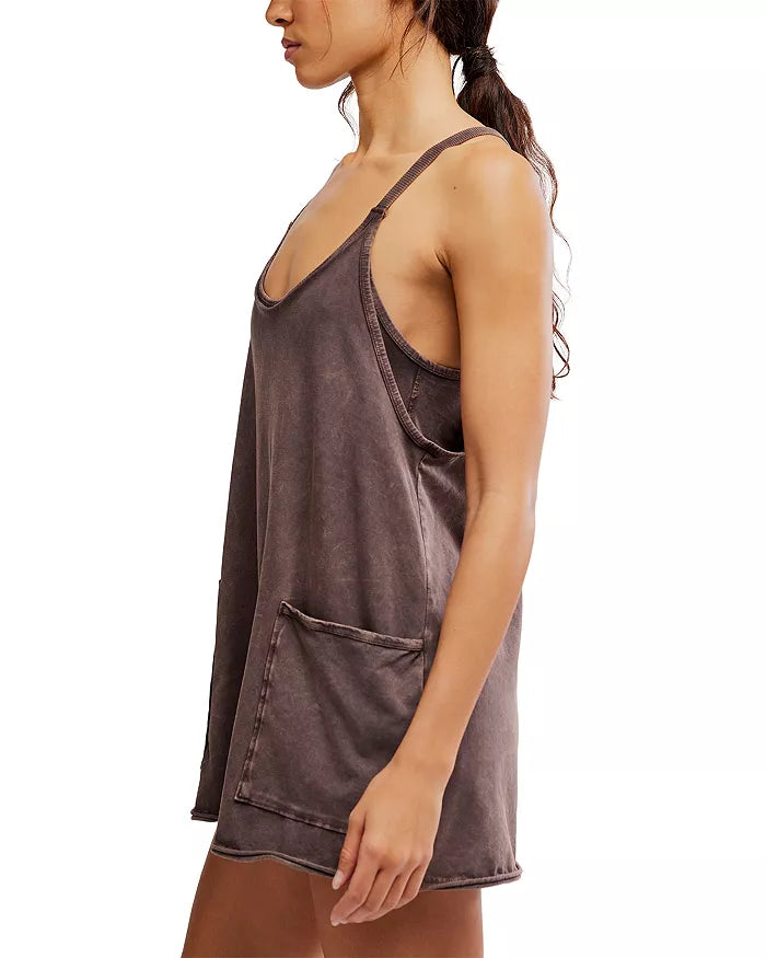 A woman stands in profile wearing the Hot Shot Mini by FP Movement—a dark brown, mini-length tank dress with a relaxed fit, thin straps, and a large side pocket. Made from cotton elastane, it pairs effortlessly with her ponytail against a white background.