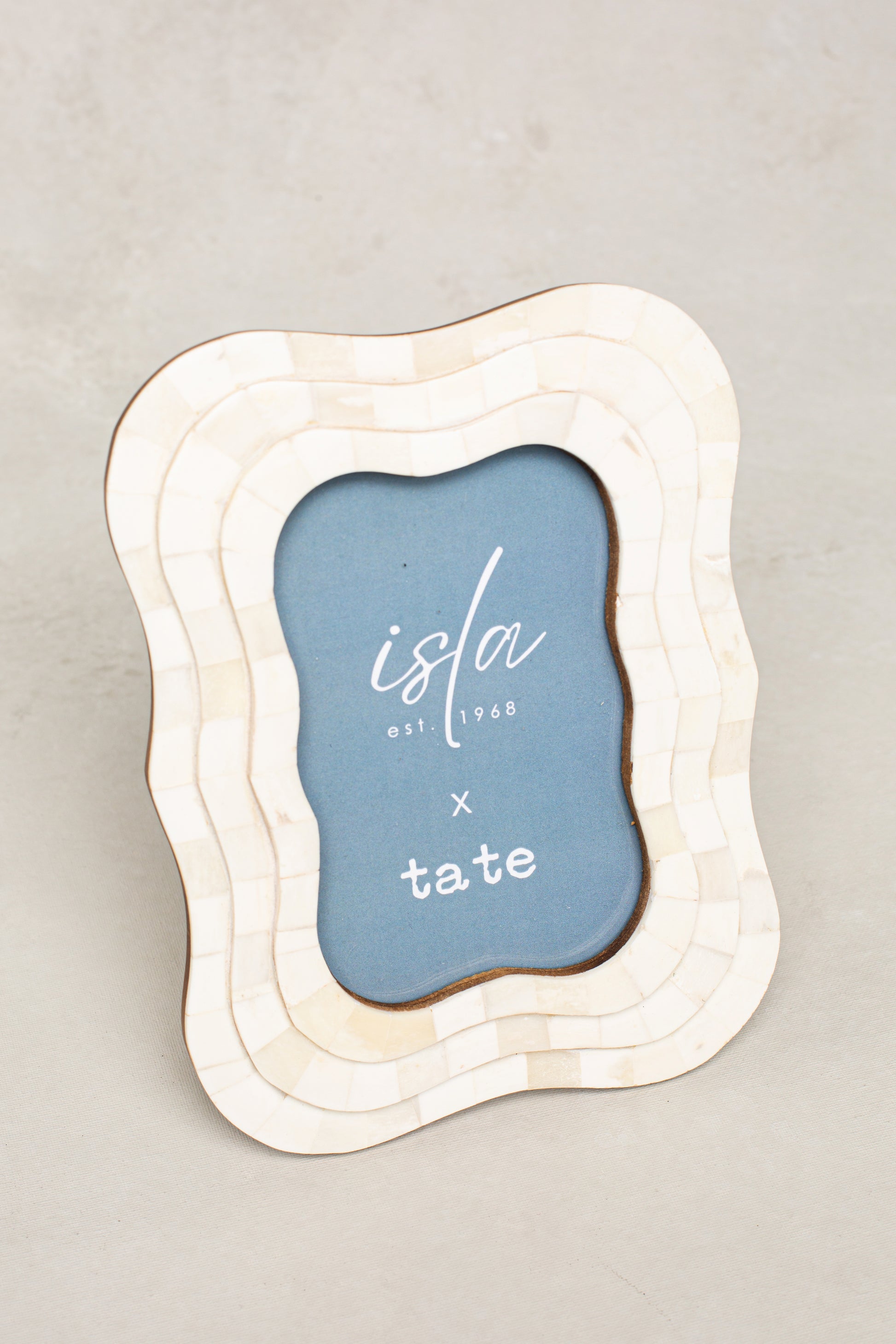 The Roma 4x6 Triple Wavy Picture Frame by Isla x Tate features an ivory handcrafted design with a blue insert reading "isla est. 1968 x tate" in white, displayed on a light gray surface.