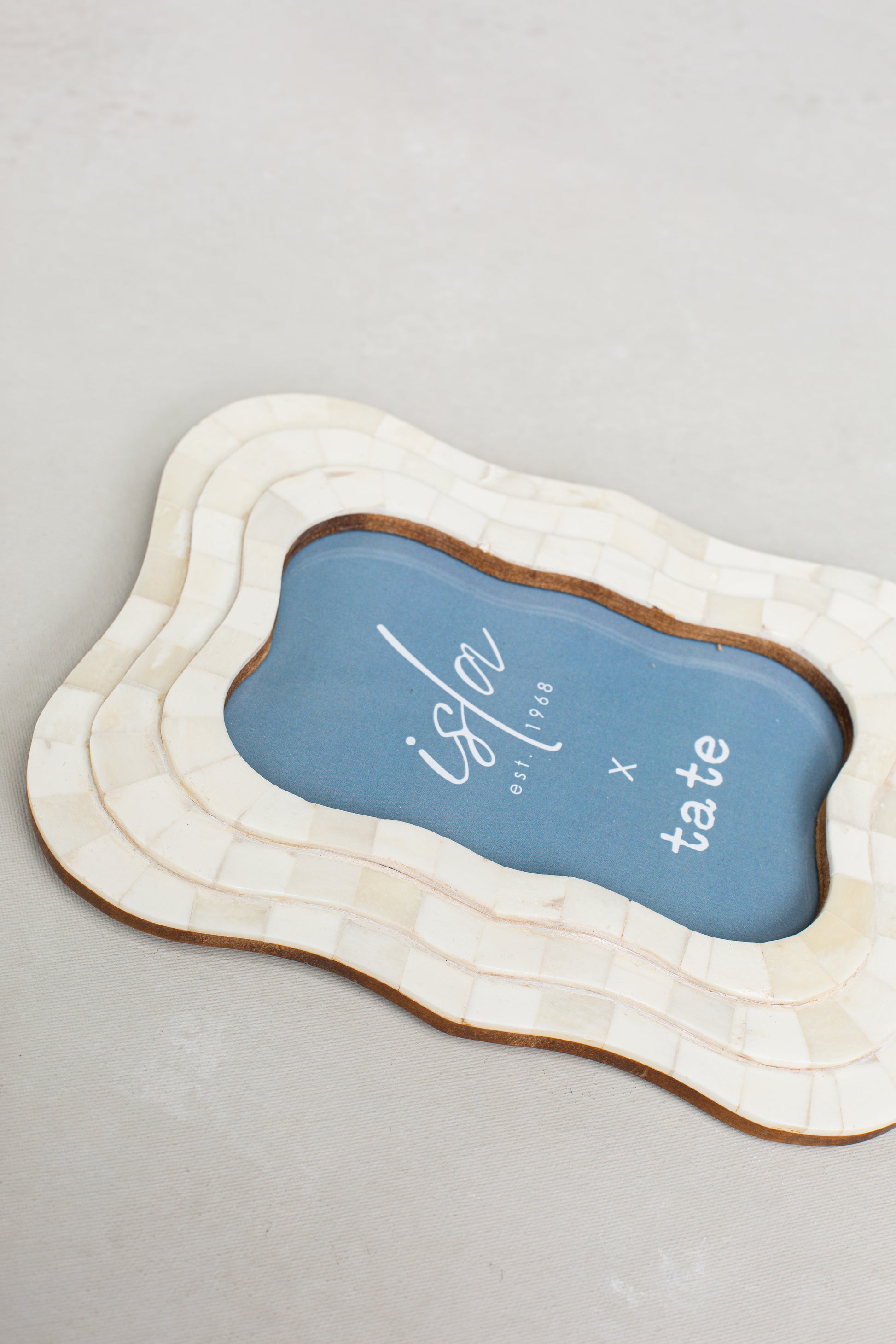 The Roma 4x6 Triple Wavy Picture Frame by Isla x Tate, featuring an ivory finish and blue insert with the brand’s name in white cursive and bold text, rests on a light textured surface.